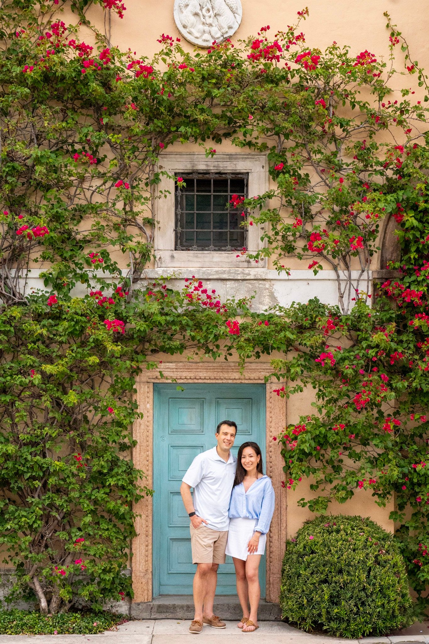 Couple poses in front of turquoise door surrounded by climbing red roses.