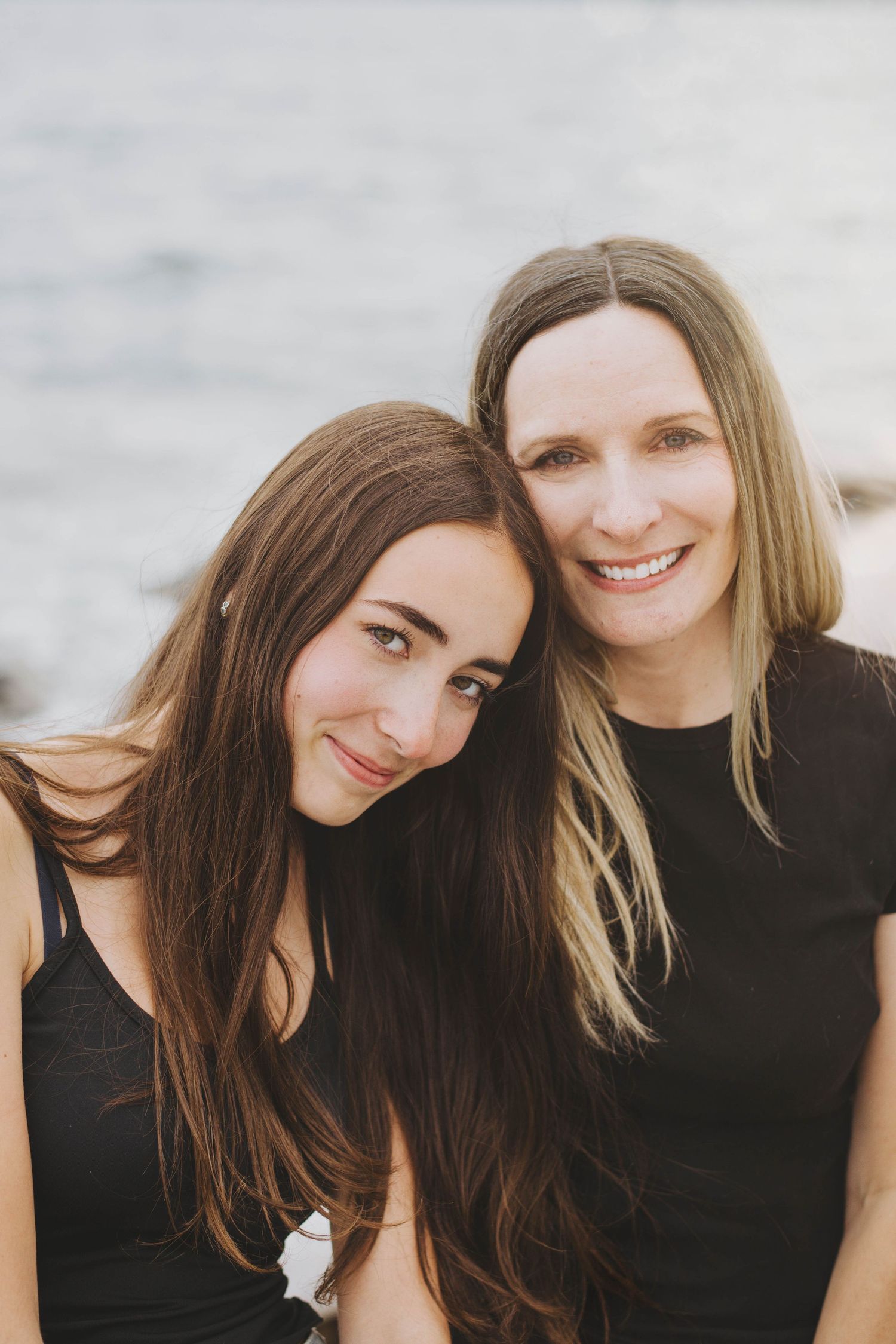 Two women with brown and blonde hair smile together outdoors near water, both wearing black tops.