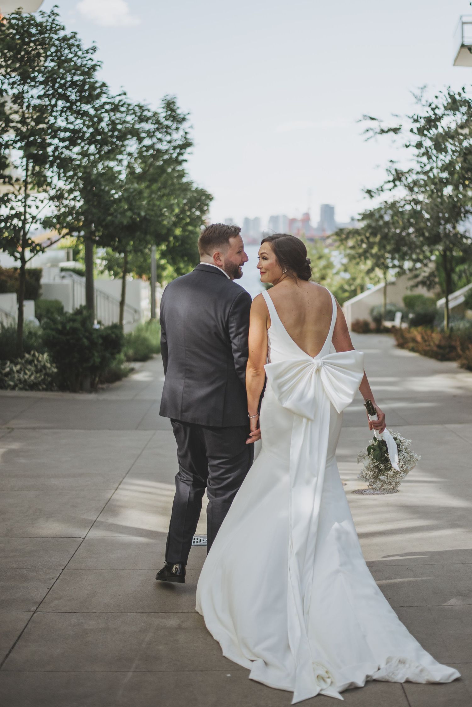 Newlyweds walk together on city path with bride wearing backless gown with large bow detail.