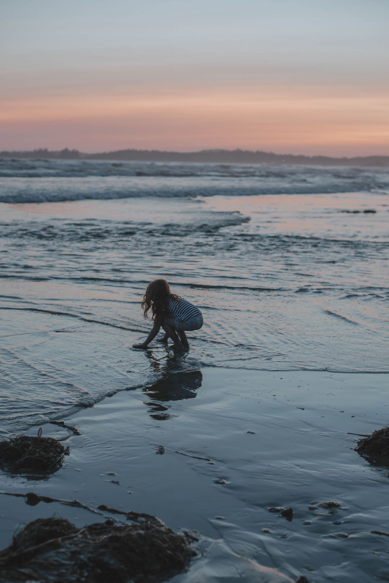 Child playing at the ocean's edge during a colorful sunset, crouching on wet sand with gentle waves rolling in.