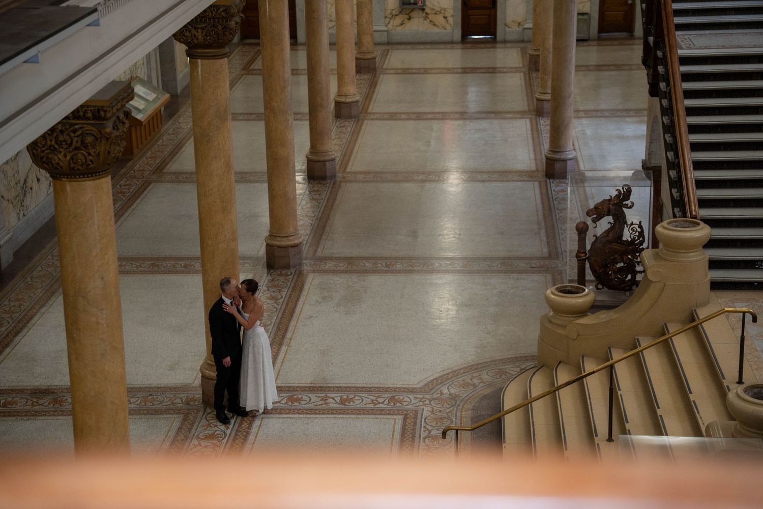 Bride and groom share a romantic moment in an elegant marble hall with tall columns and grand staircase.