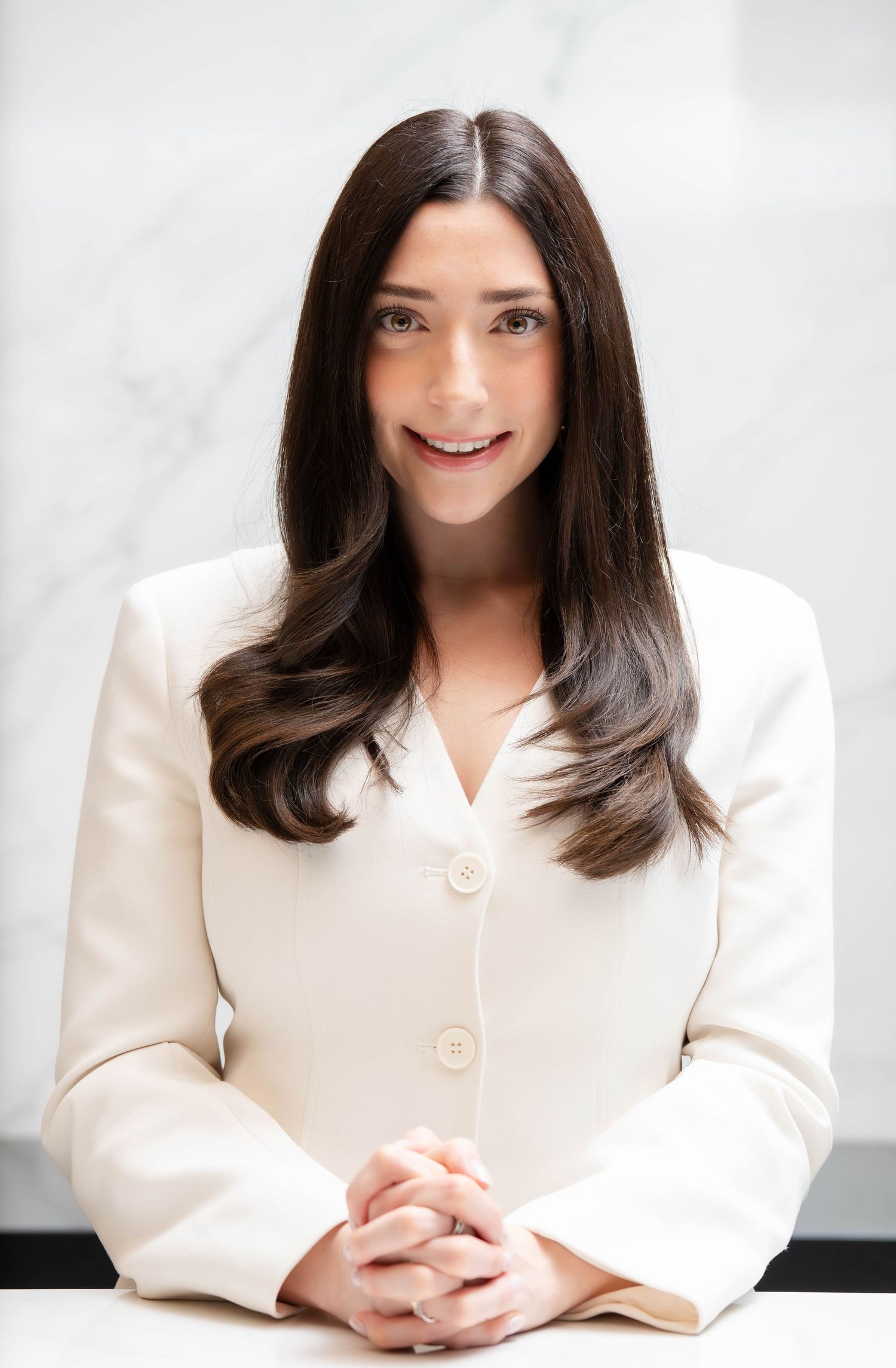 Professional woman in cream blazer with long dark hair, smiling confidently with hands clasped at a marble desk.