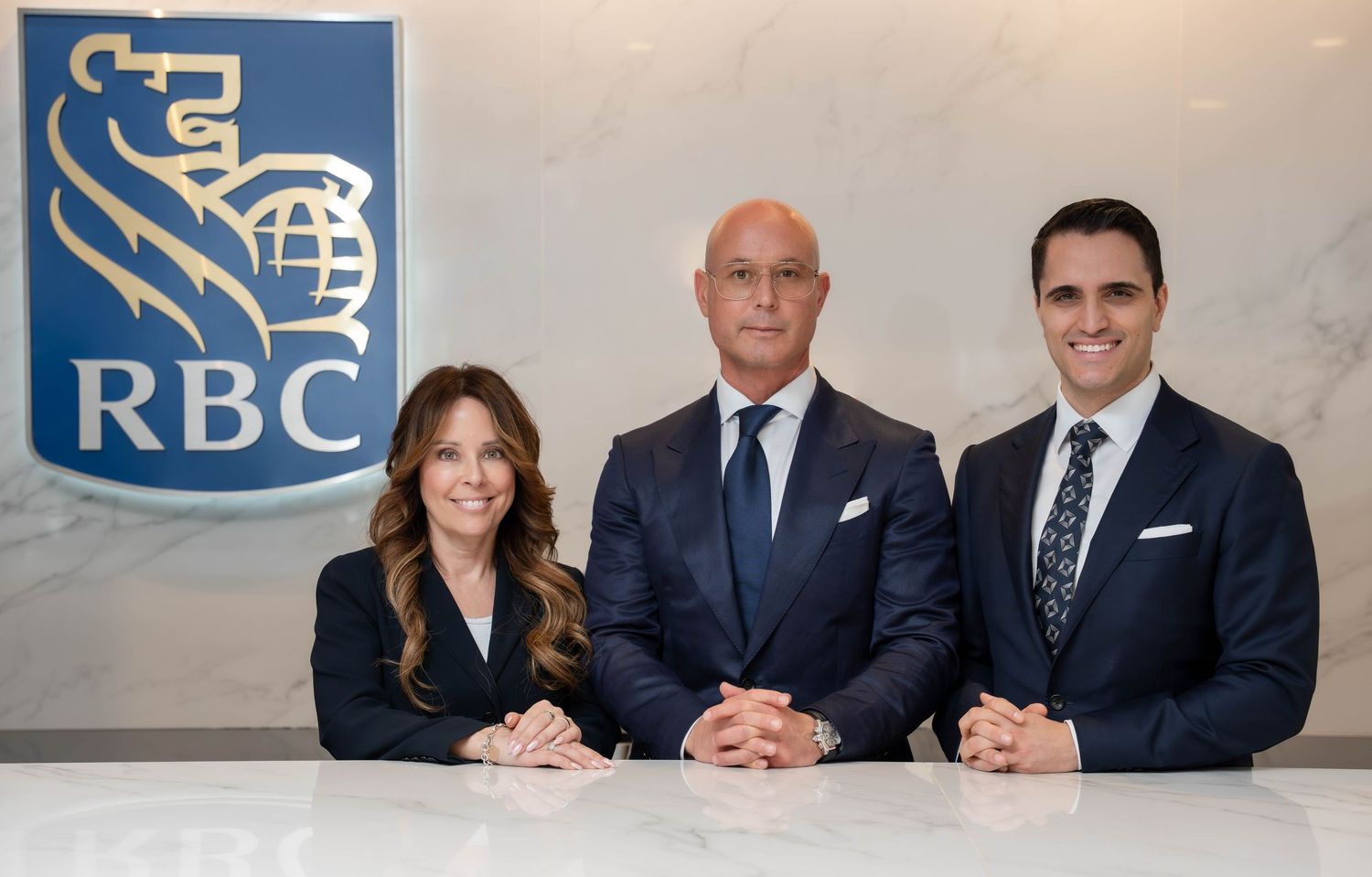 Three RBC financial professionals in formal attire posing together in front of an RBC branded office backdrop.