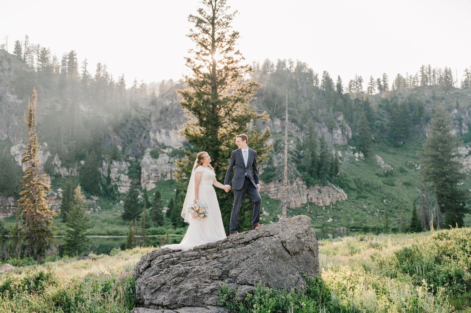 Newlyweds standing on rock formation surrounded by mountain scenery.