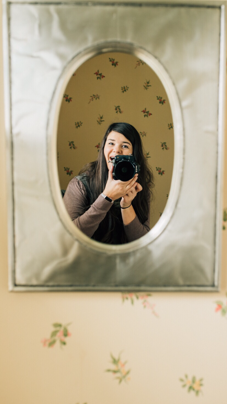 Mirror selfie showing photographer with camera against floral wallpaper background.