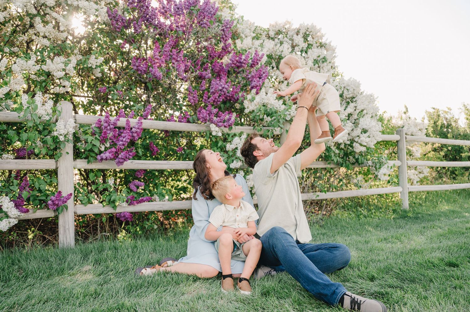 Family relaxes together on grassy hill with blooming purple flowers and white fence in background.