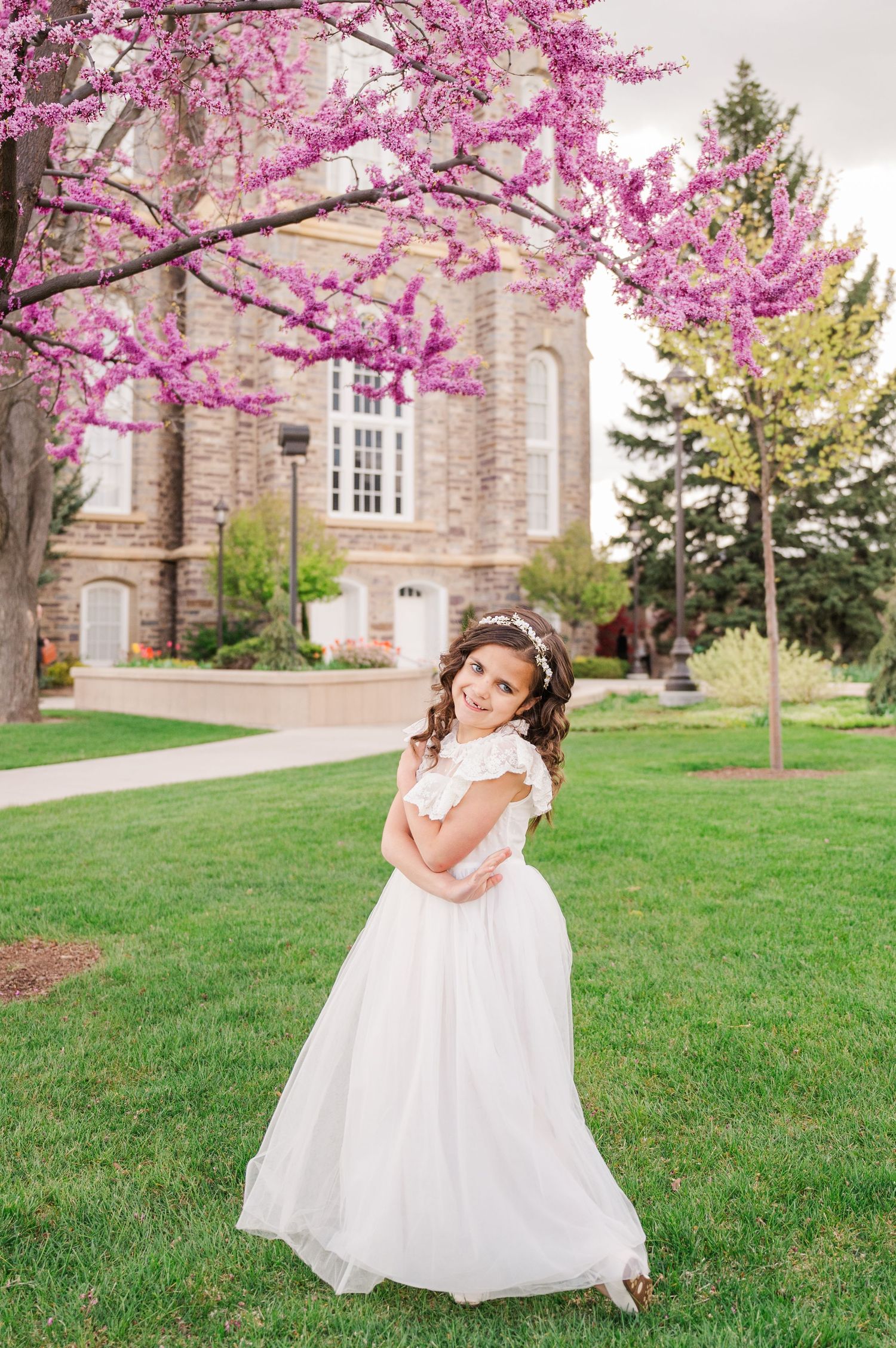 Person in white dress twirling near blooming cherry trees with historic building in background.