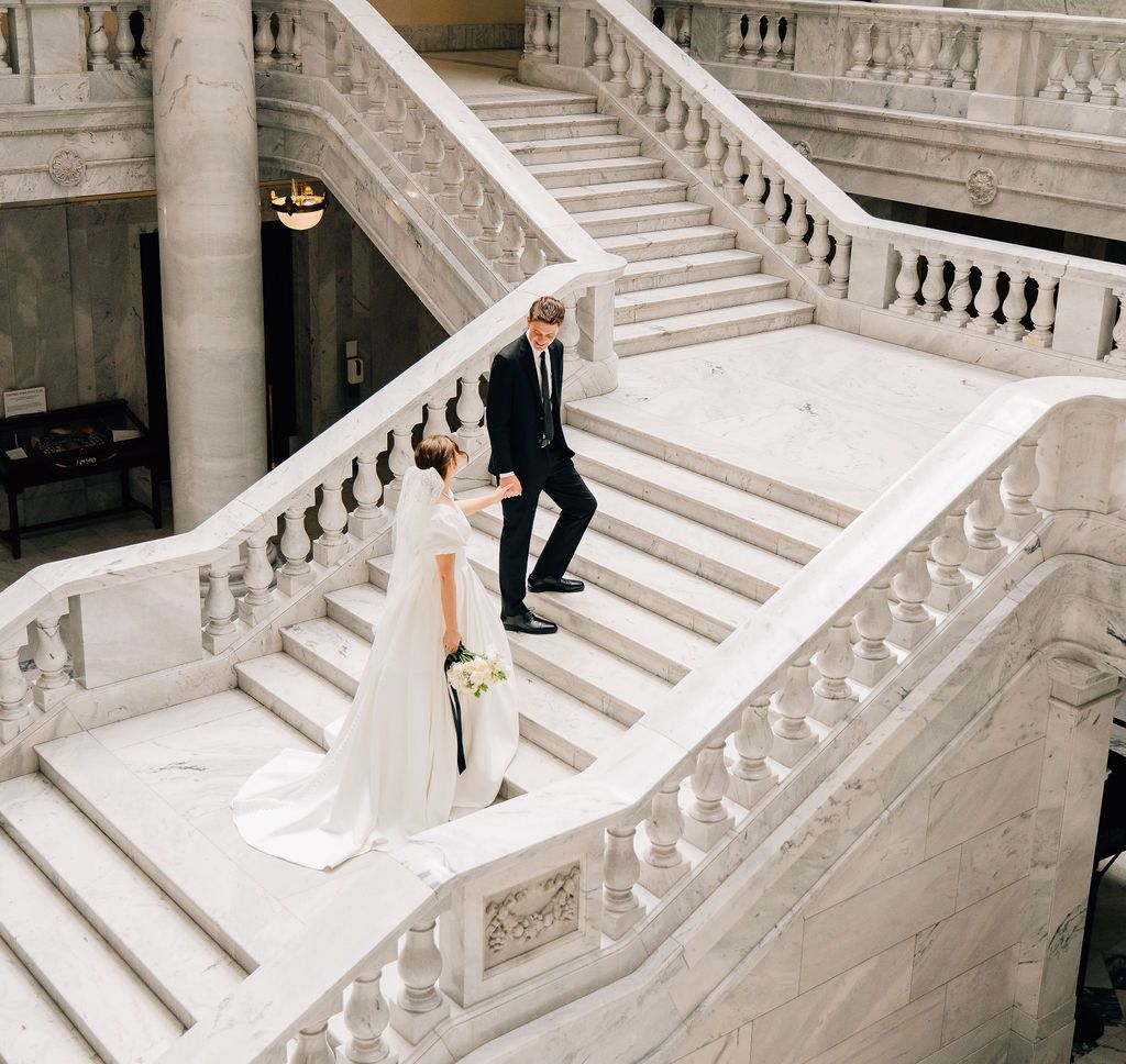 Bride and groom walking down elegant white marble staircase in grand building during wedding photo session.