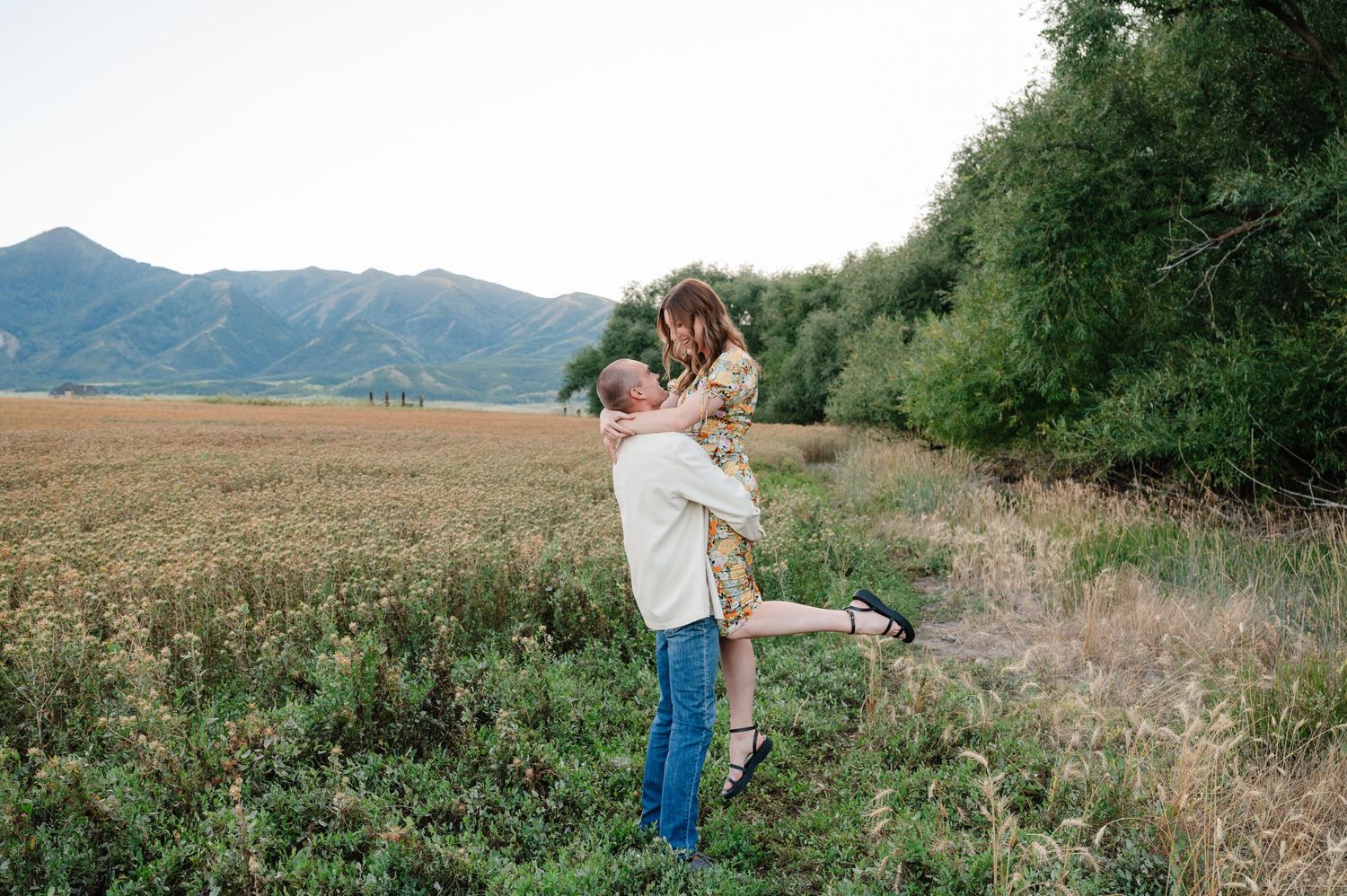 A couple embraces in a field with mountains and trees in the background during golden hour.