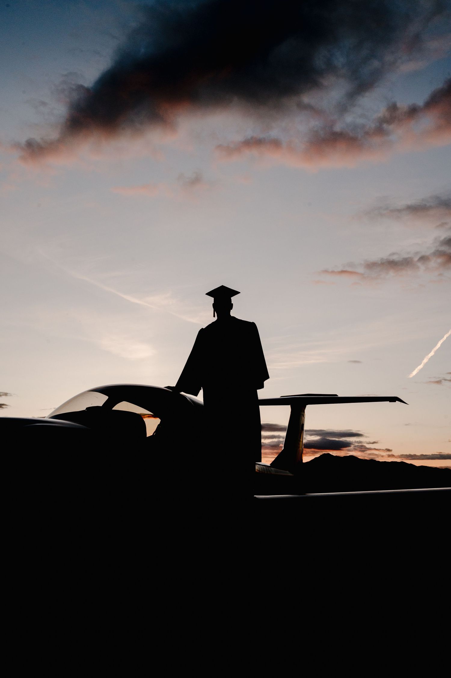 Silhouette of a graduate in cap and gown stands beside a car against a dramatic sunset sky with clouds.