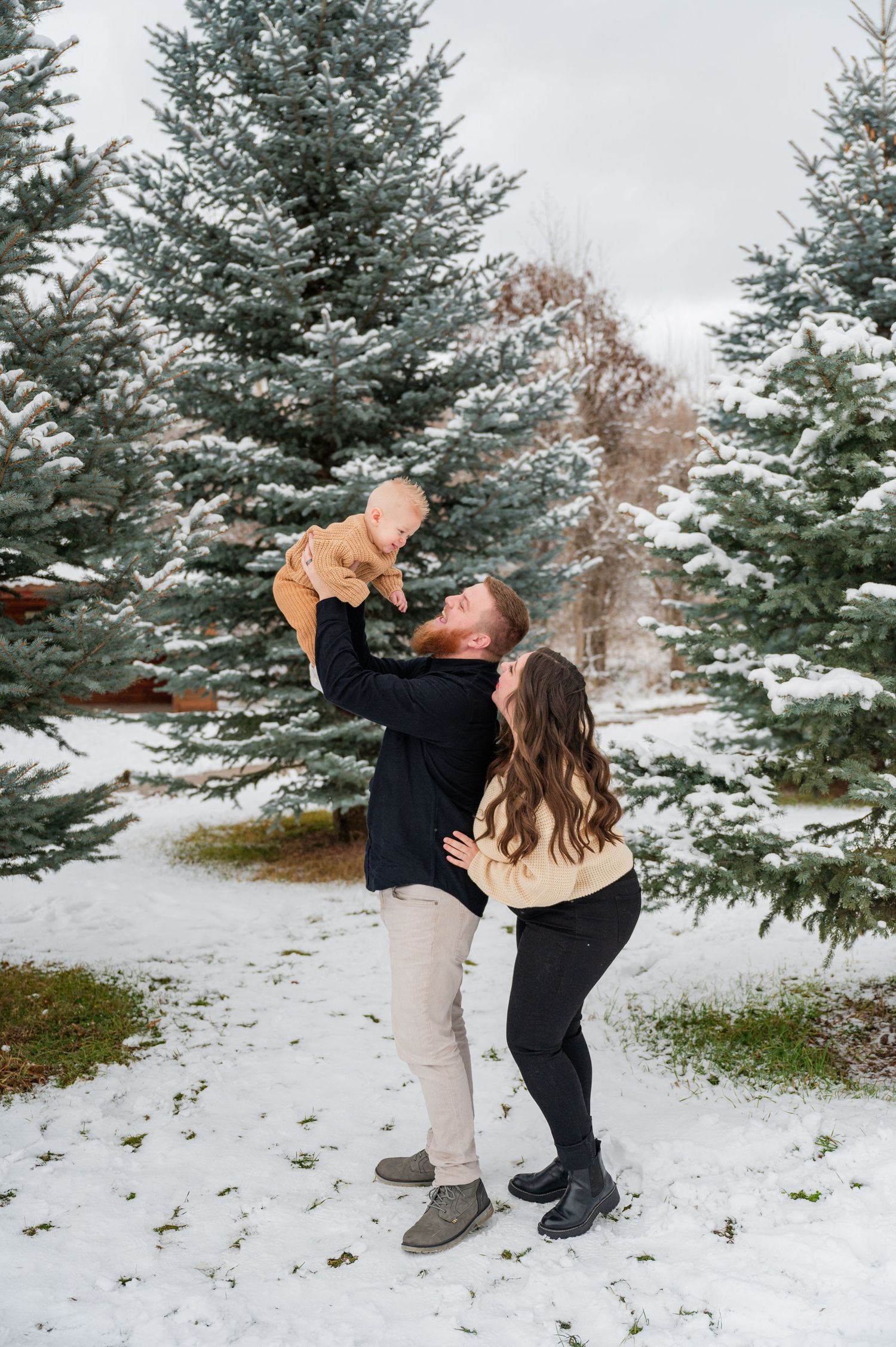 A couple shares a playful moment lifting their baby in a snowy evergreen forest.