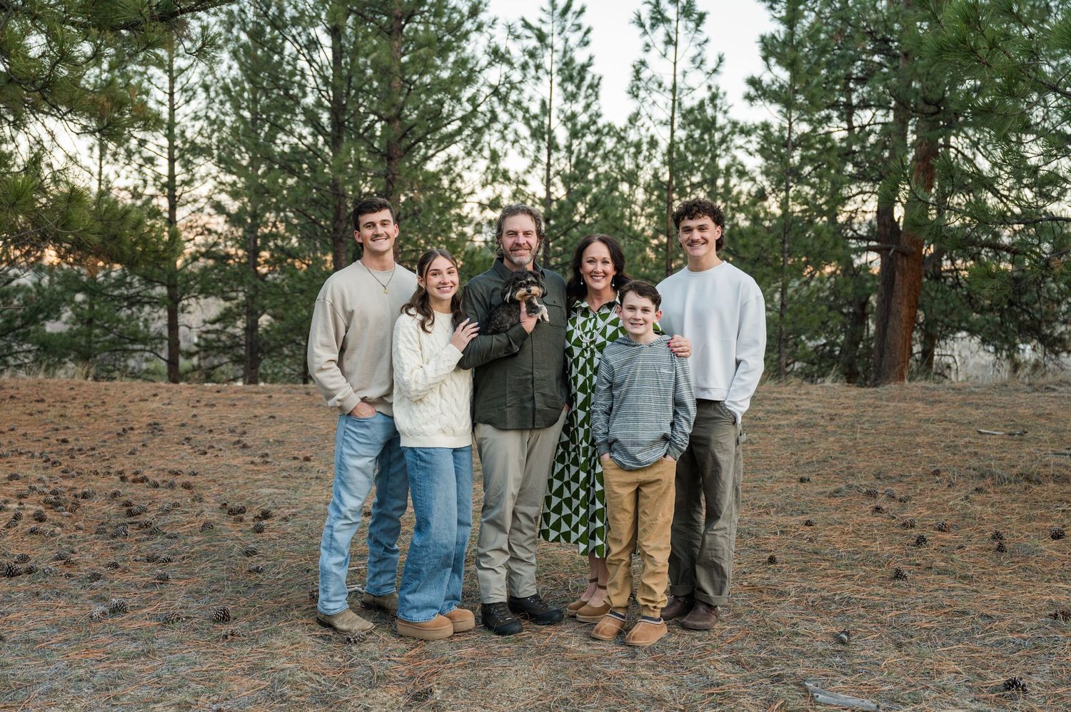 A casual group portrait of people wearing casual clothing standing together outdoors among pine trees.