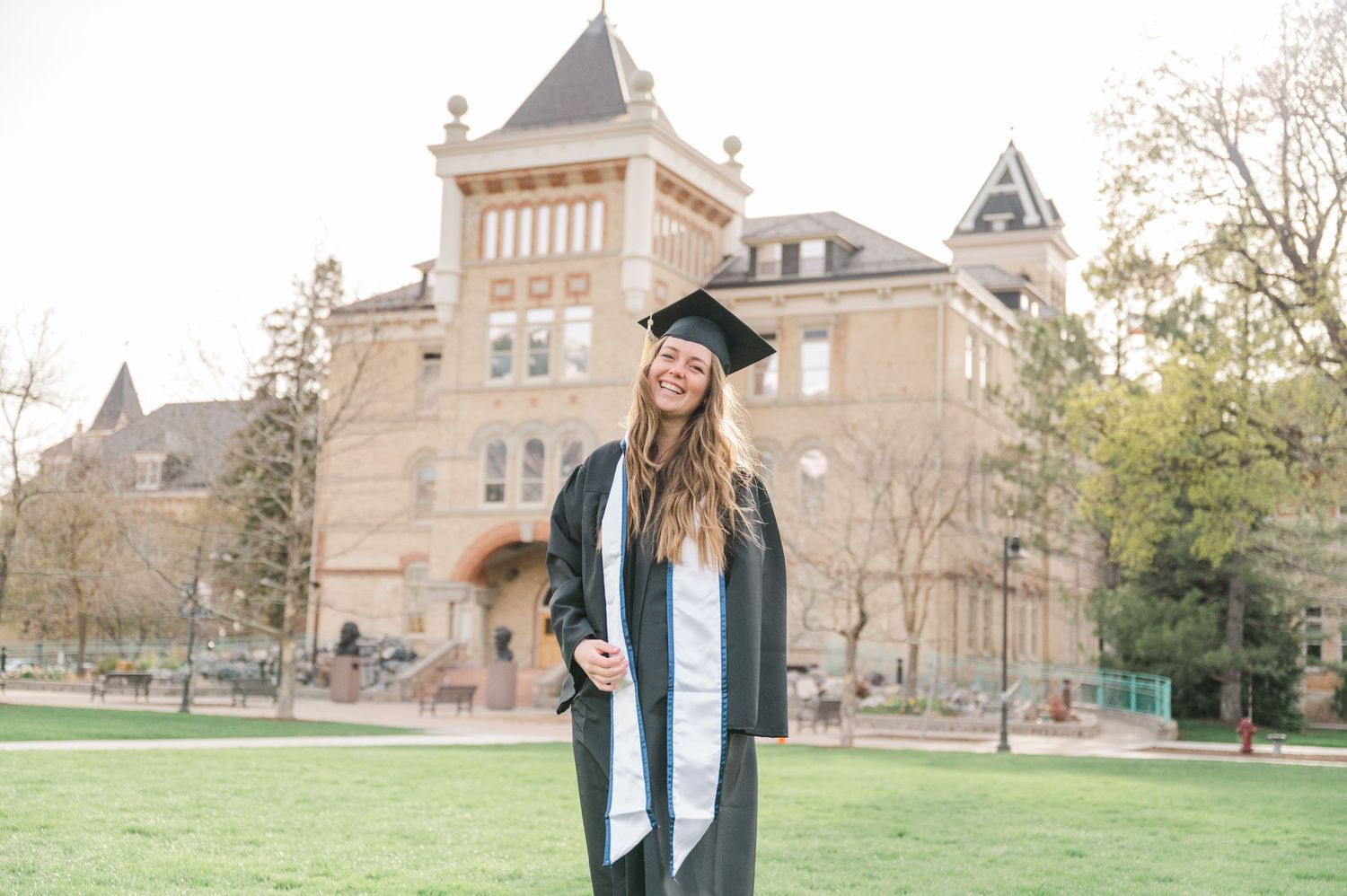 Graduate in cap and gown poses in front of historic university building on sunny day.