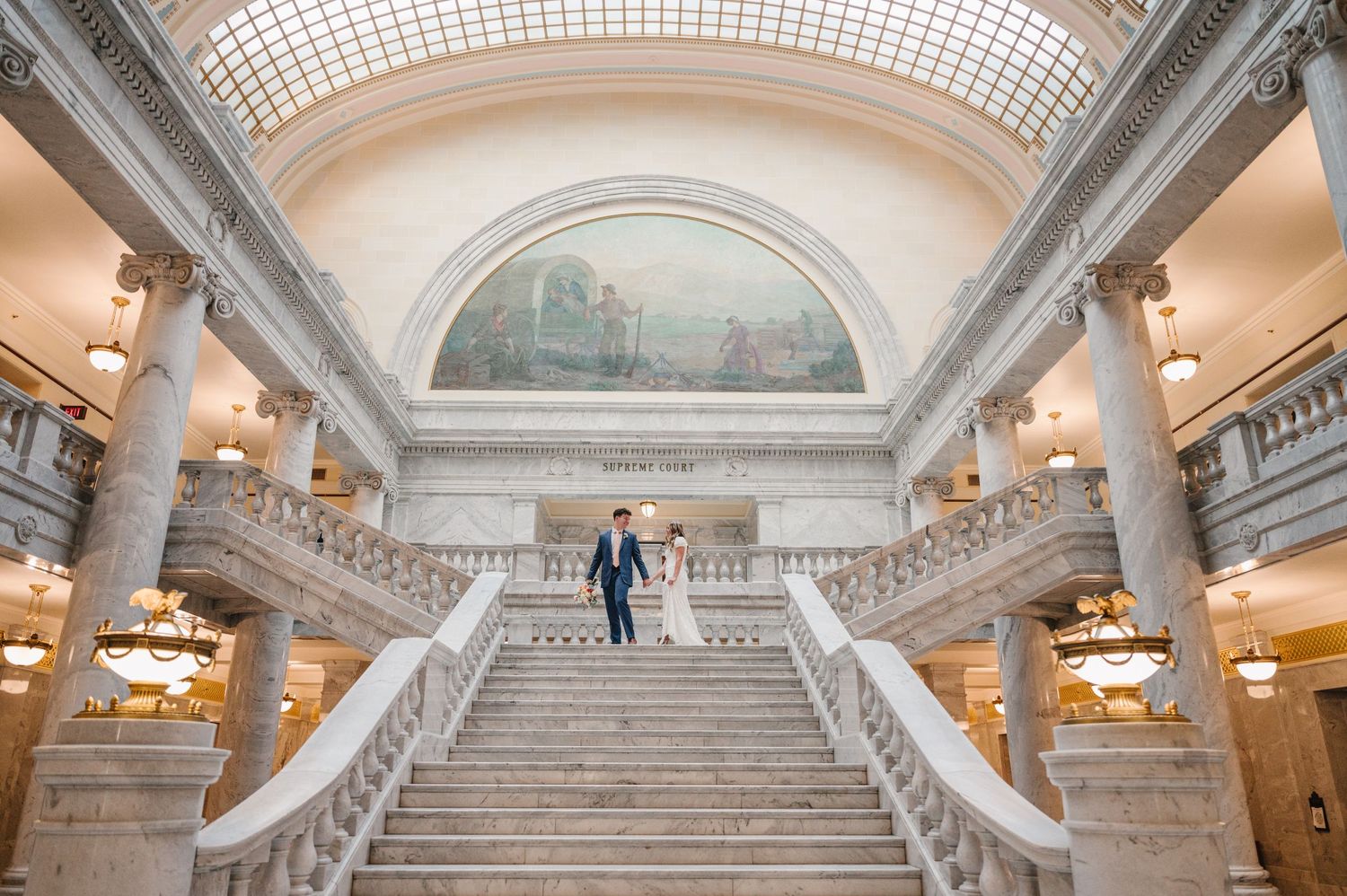 Couple stands on ornate marble staircase in historic building with vaulted ceiling and elegant lighting.
