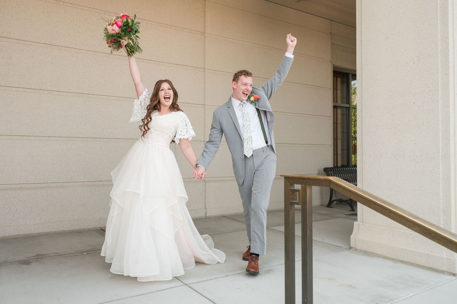 Newlyweds celebrate with raised arms while walking down an outdoor corridor.