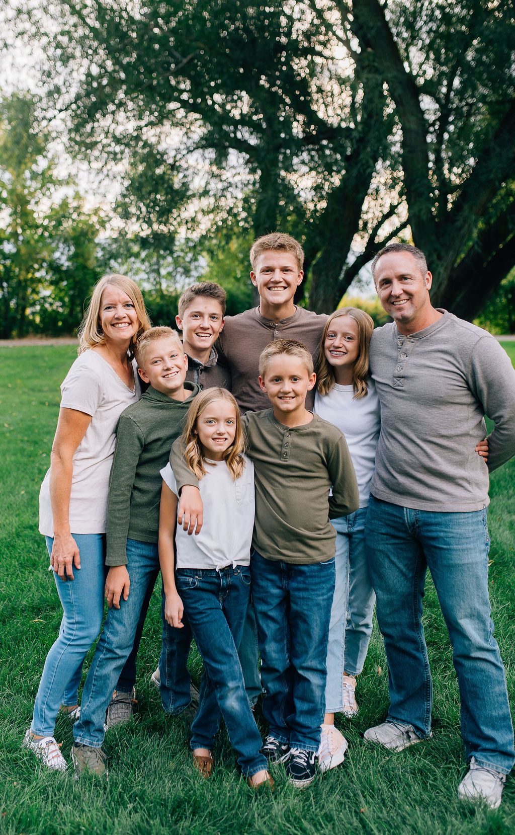 A family of seven stands together outdoors on grass with trees in the background during a casual photo session.