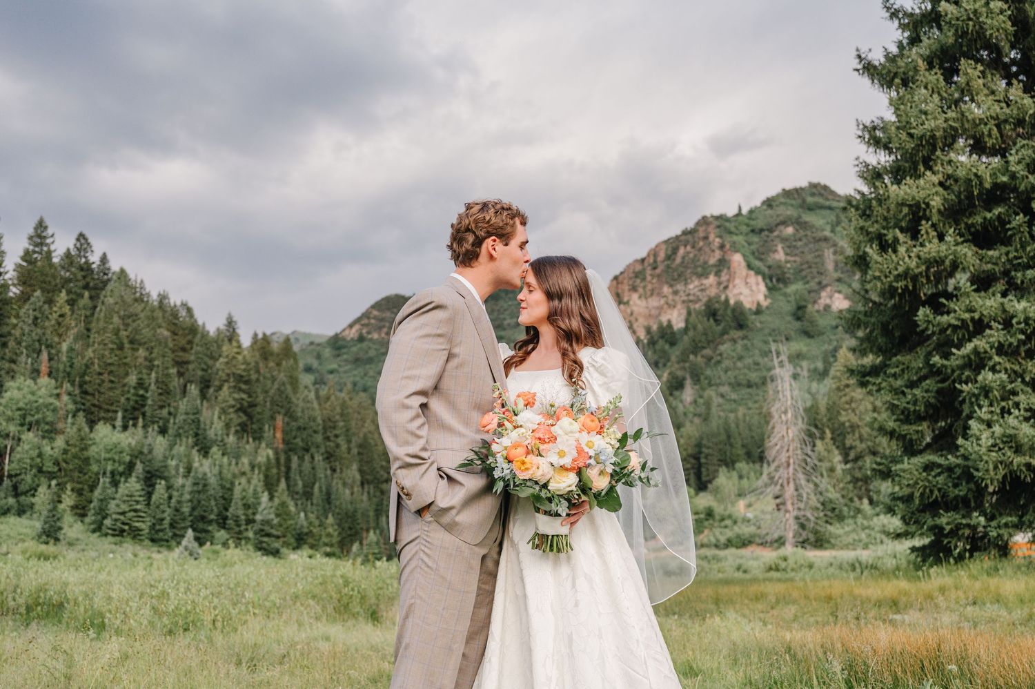 A couple shares a romantic moment in a scenic mountain meadow surrounded by evergreen trees and wildflowers.