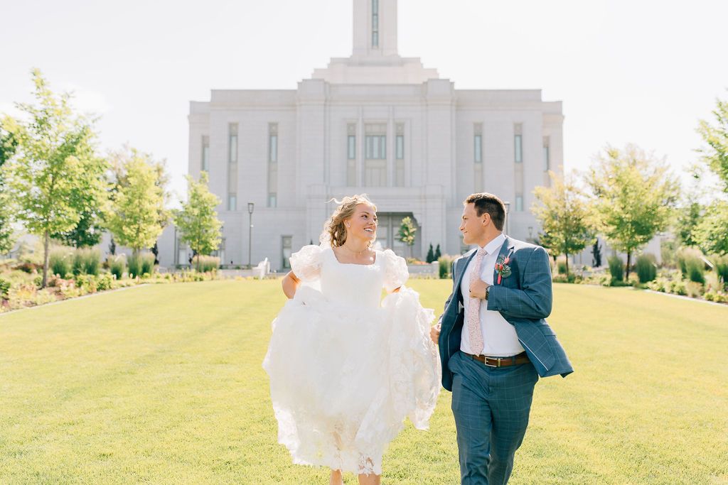 A couple in wedding attire dances and twirls together on a sunny grassy lawn with a white building in the background.