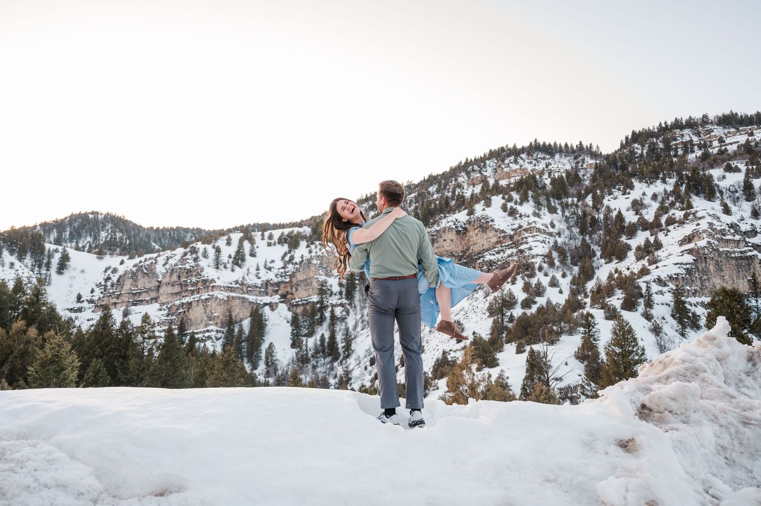 A couple walks together on a snowy mountain trail with scenic winter peaks in the background.