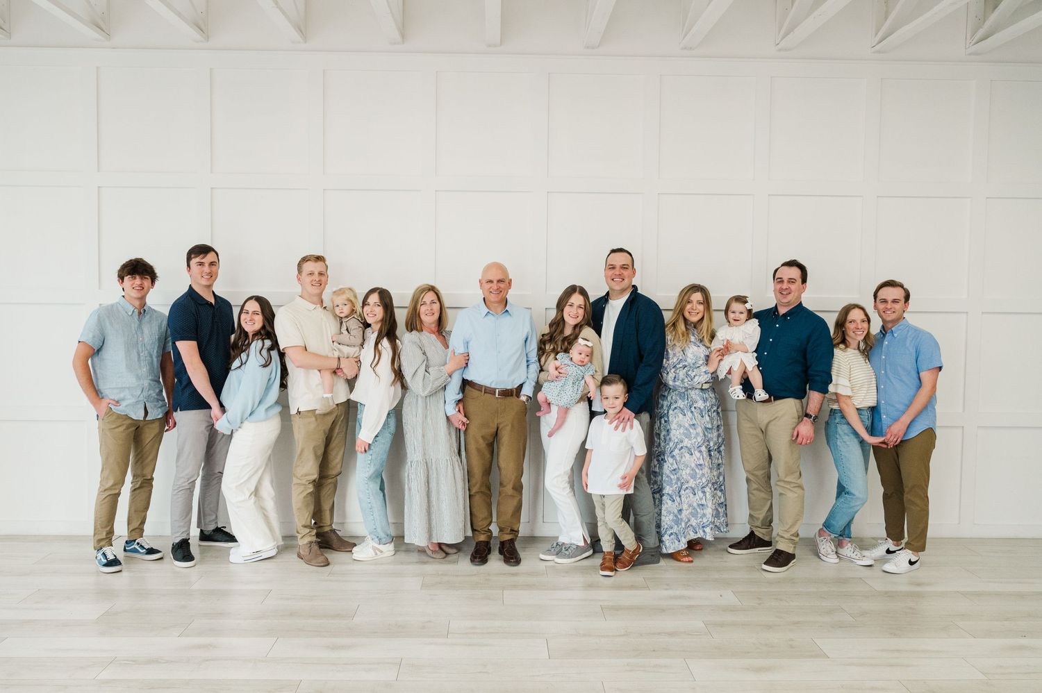Large extended family group photo against white wall wearing coordinated blue and neutral tones.