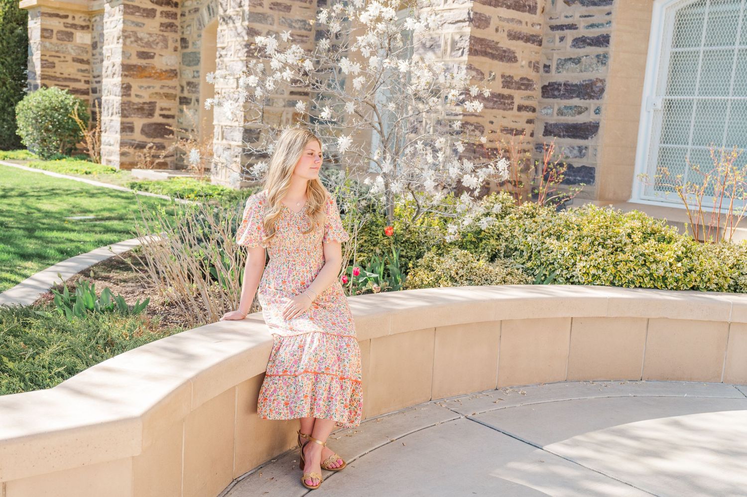 Person in floral dress sitting on stone ledge outside building with landscaping.