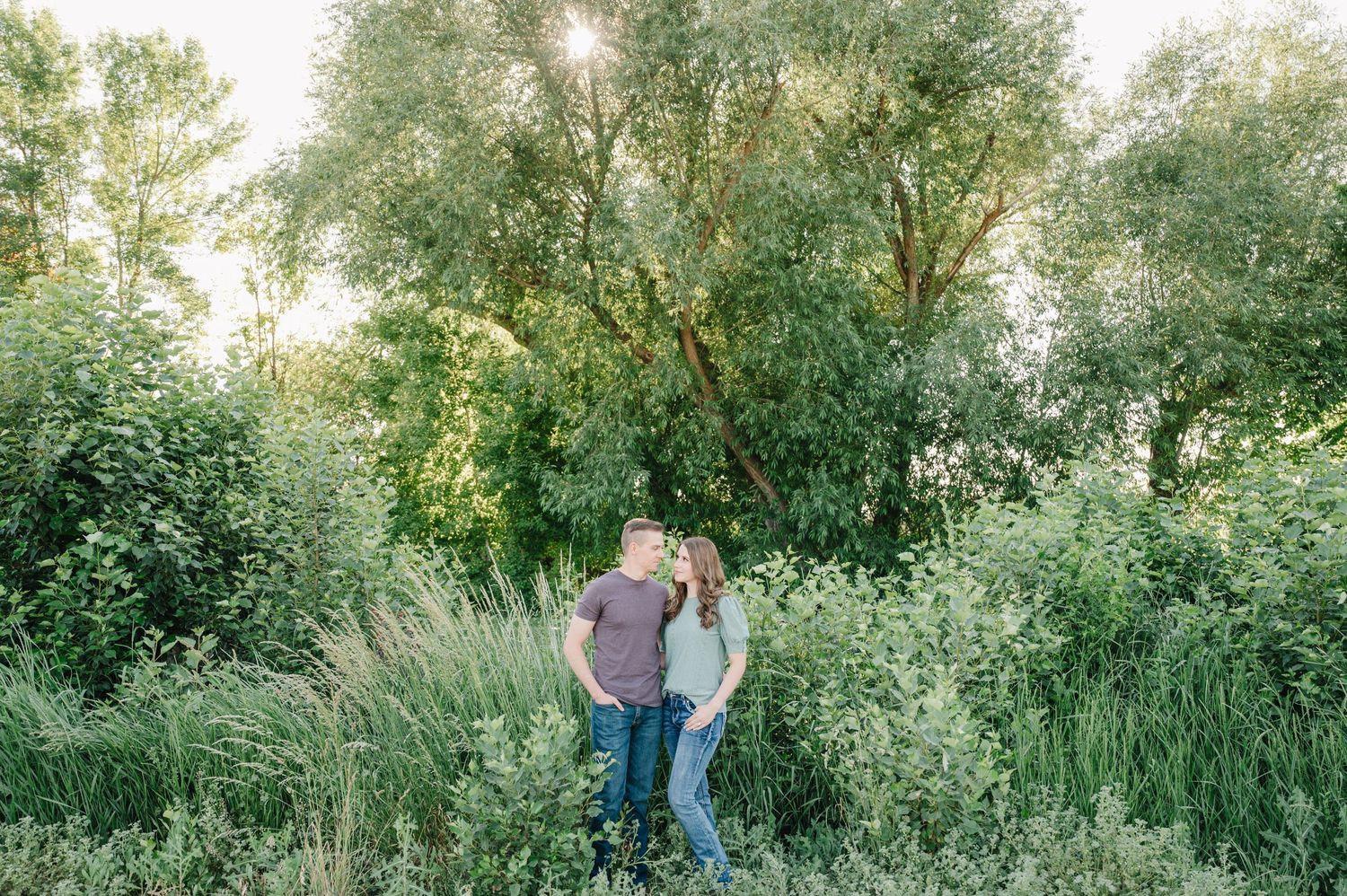 A couple sharing an intimate moment among lush green trees and tall grass at sunset.