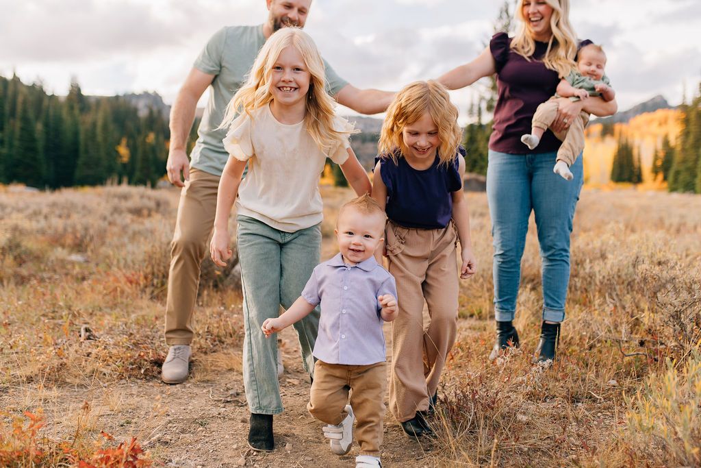 Family walking together through golden autumn field at sunset while holding hands and smiling.