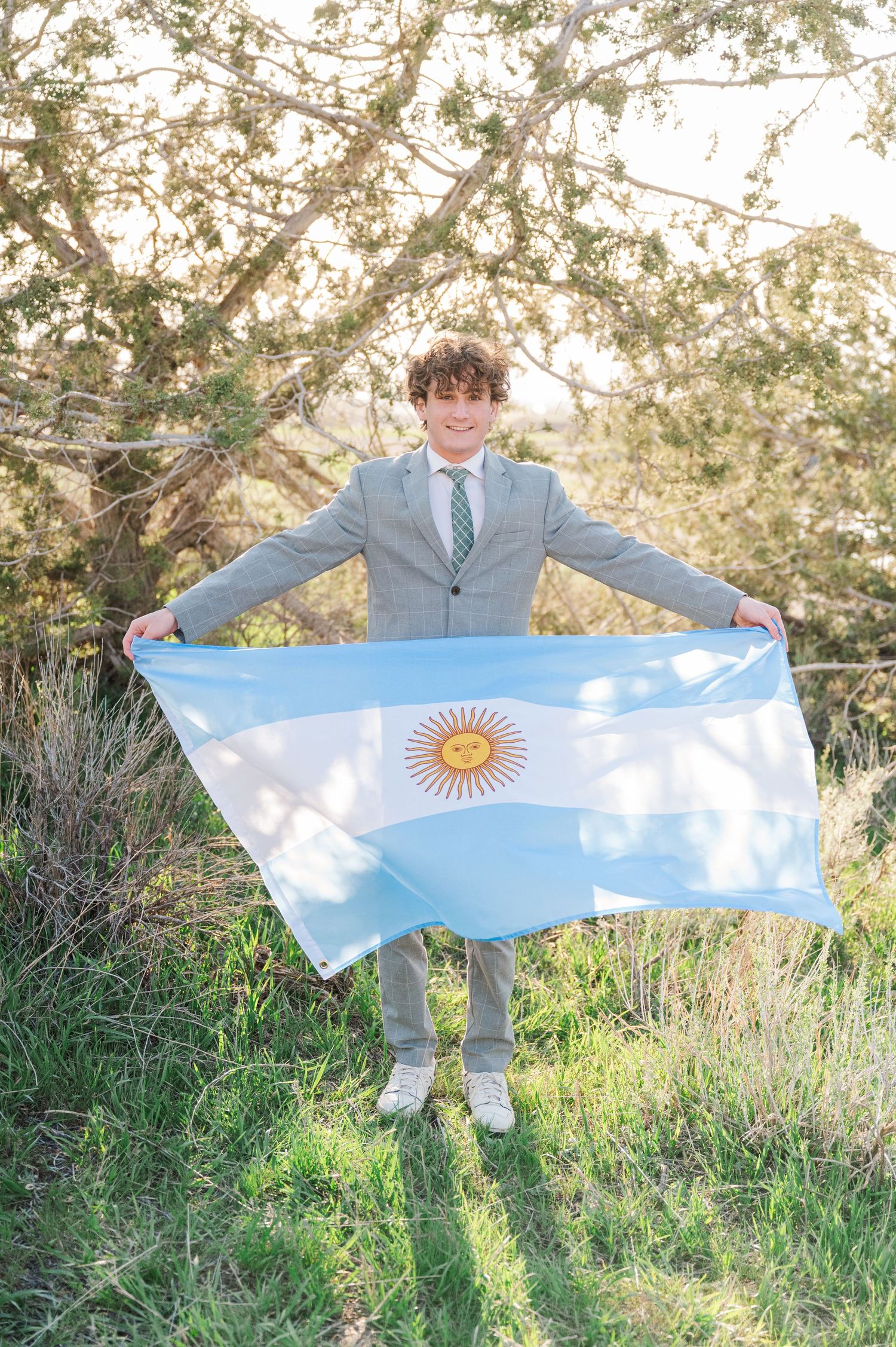 Person in light gray suit holding Argentine flag outdoors with trees in background.