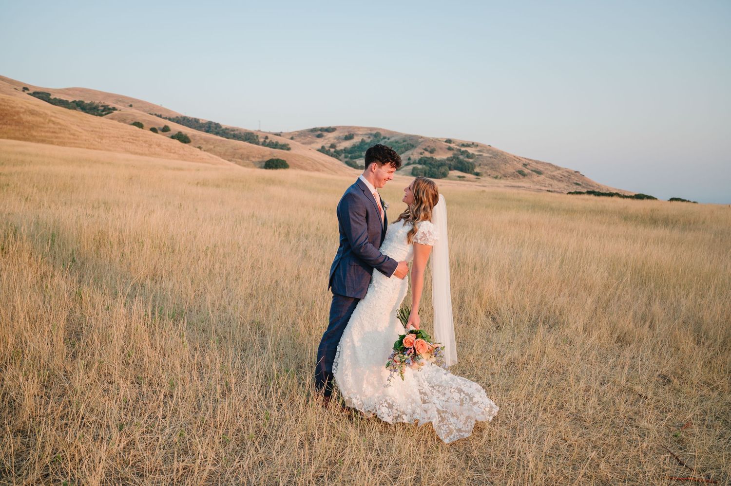 Wedding couple embracing in golden hillside field at sunset.