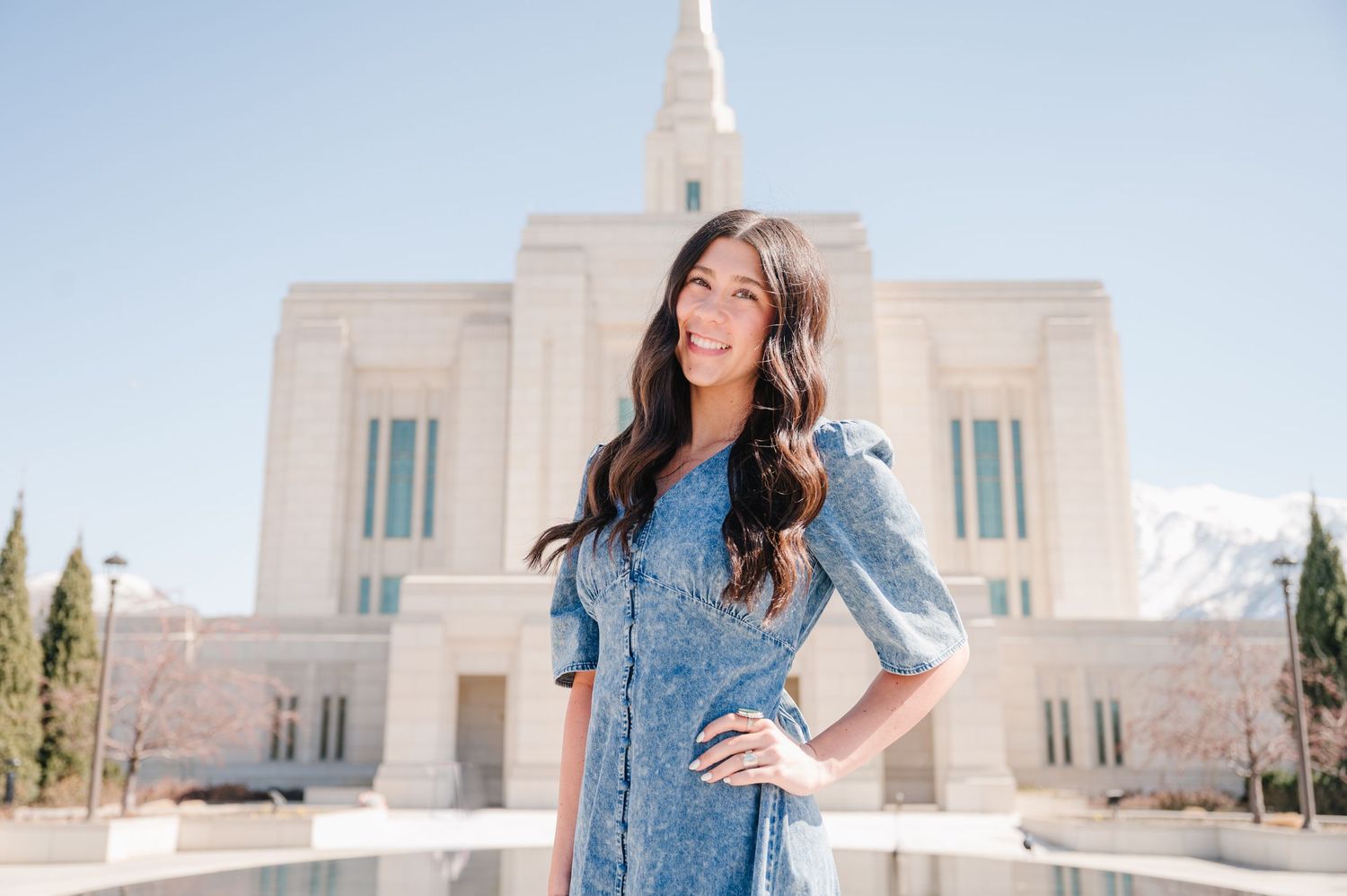 Someone in a light blue dress poses in front of a bright white Mormon temple building under a clear blue sky.