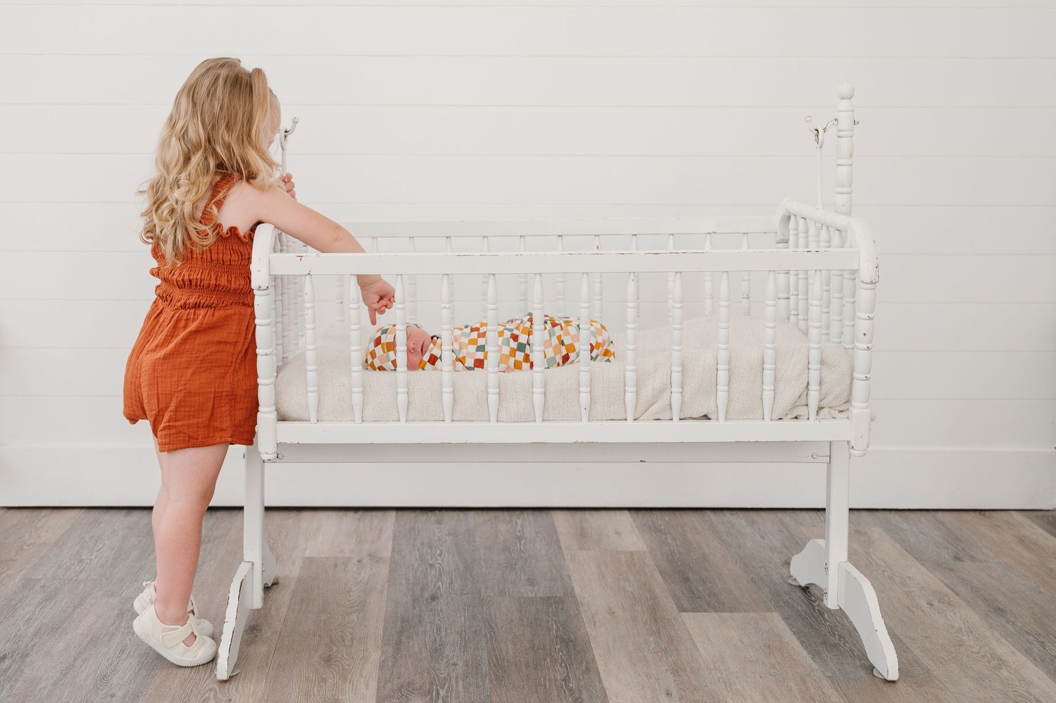 Small child in orange dress standing next to white vintage style swinging baby crib with floral decorations.