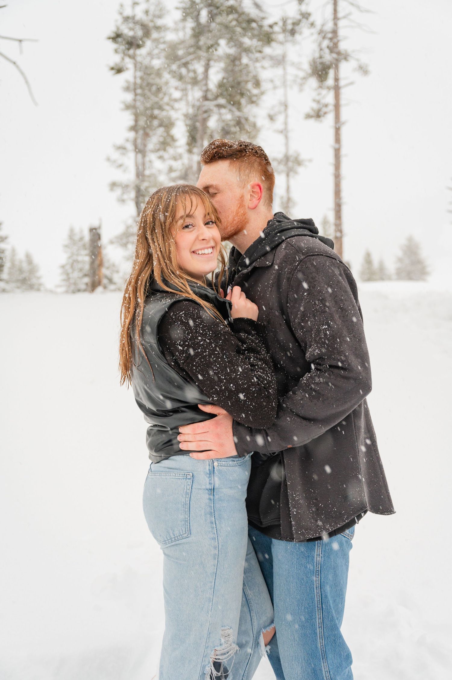 A couple embraces in a snowy forest setting while snowflakes fall around them.