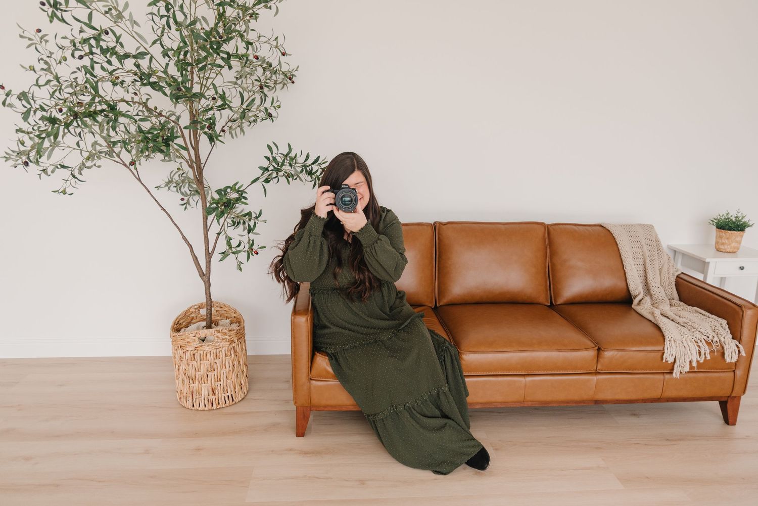 A person in a long green dress lounges on a brown leather couch next to a potted tree in a minimalist room.