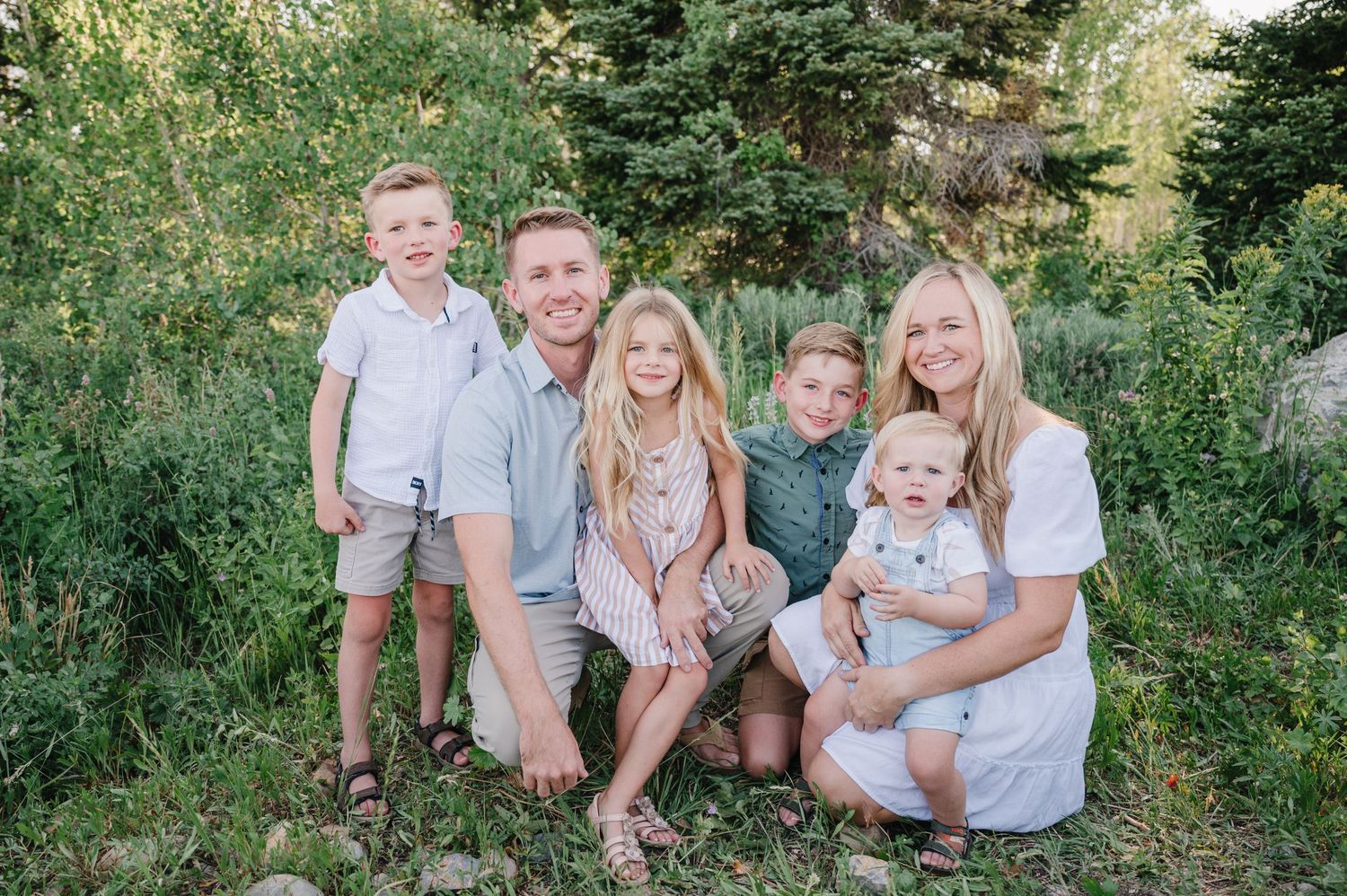 Family of six posing together on rocks amid evergreen trees in coordinated casual outfits.
