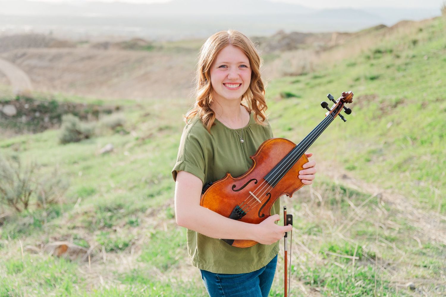 Young musician poses with violin on sunny hillside with scenic valley backdrop.
