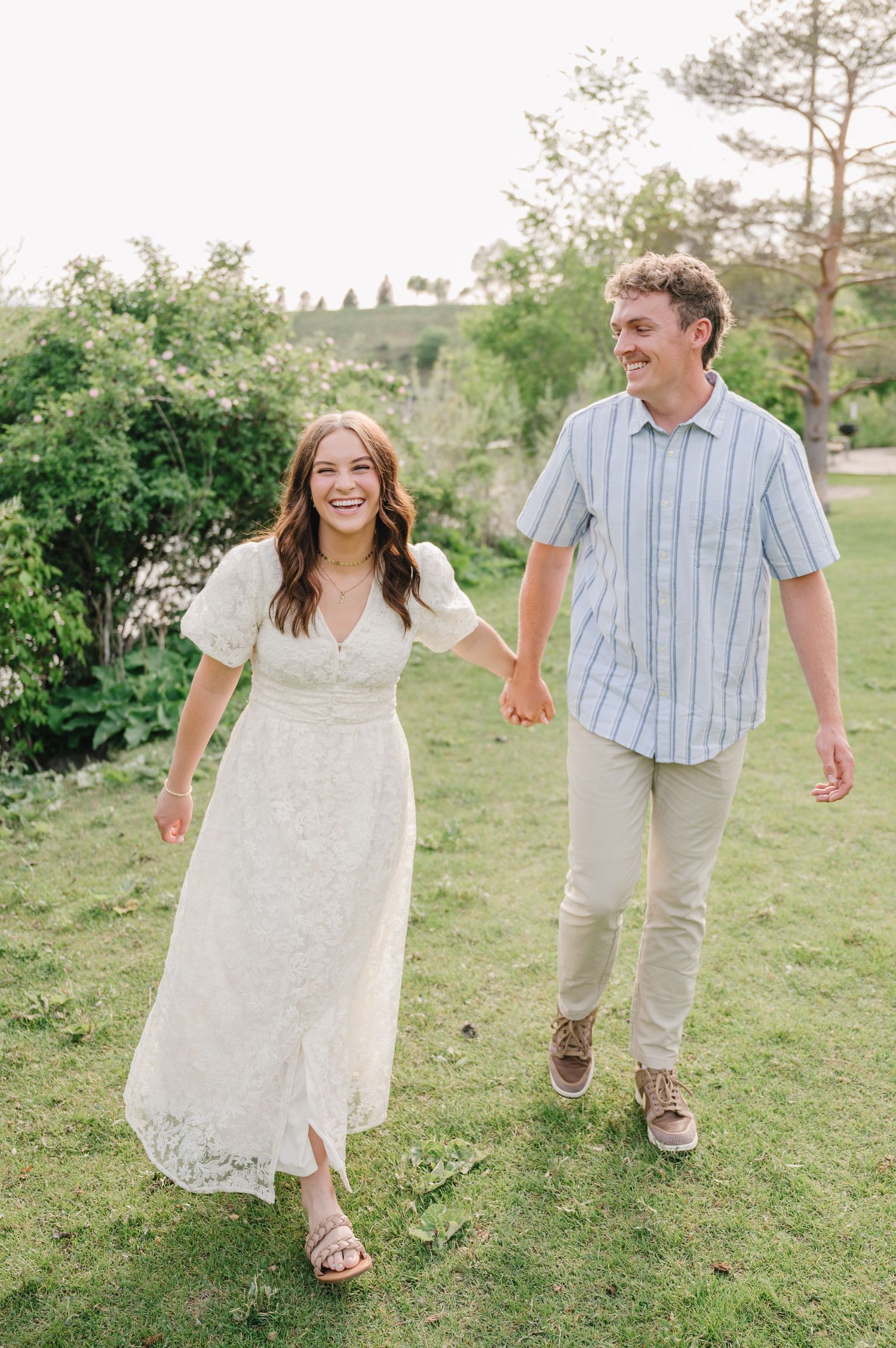 A couple walking together and laughing on a grassy hillside surrounded by greenery on a sunny day.