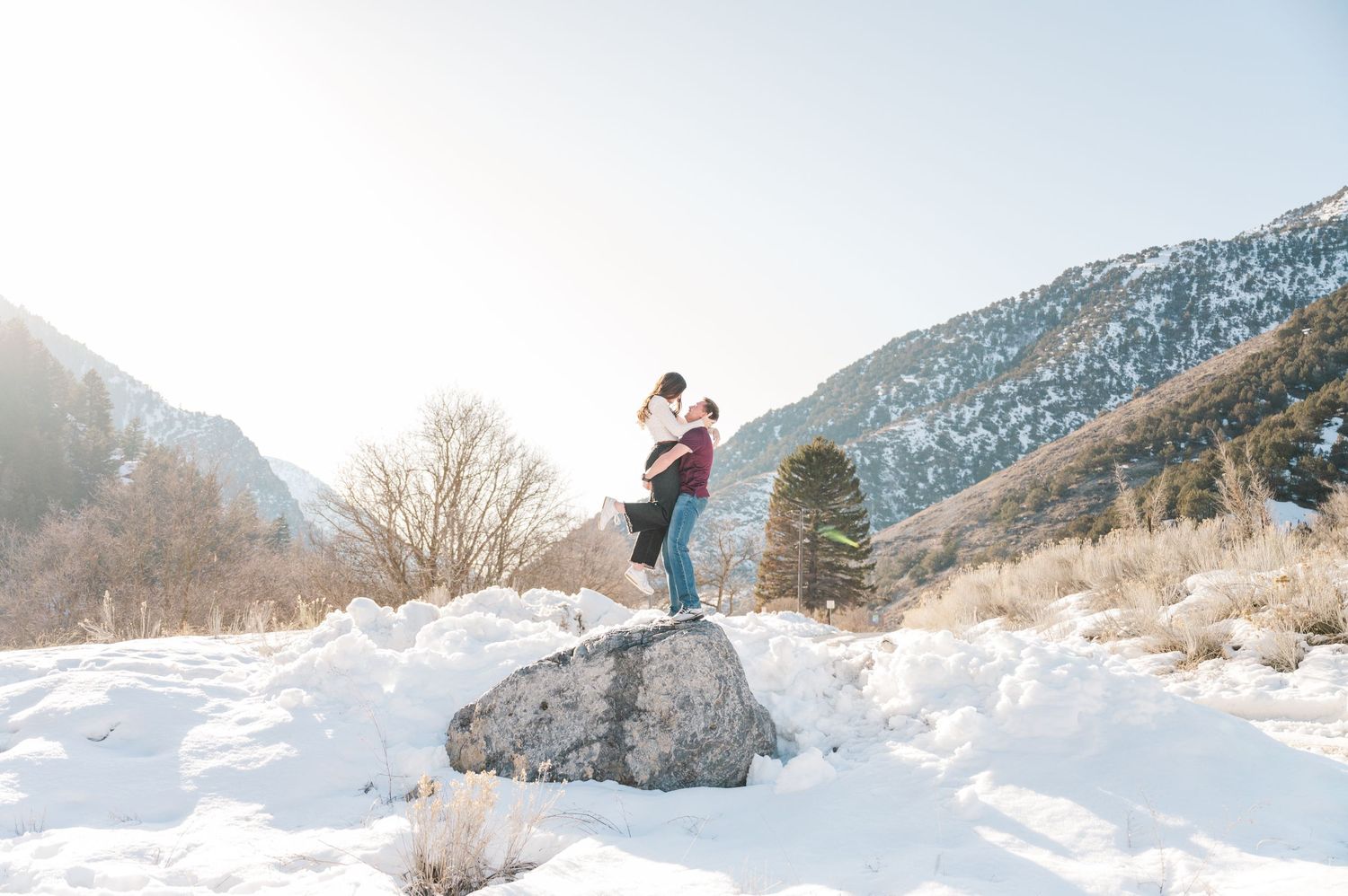 A couple embraces on a snow-covered boulder with mountain scenery.