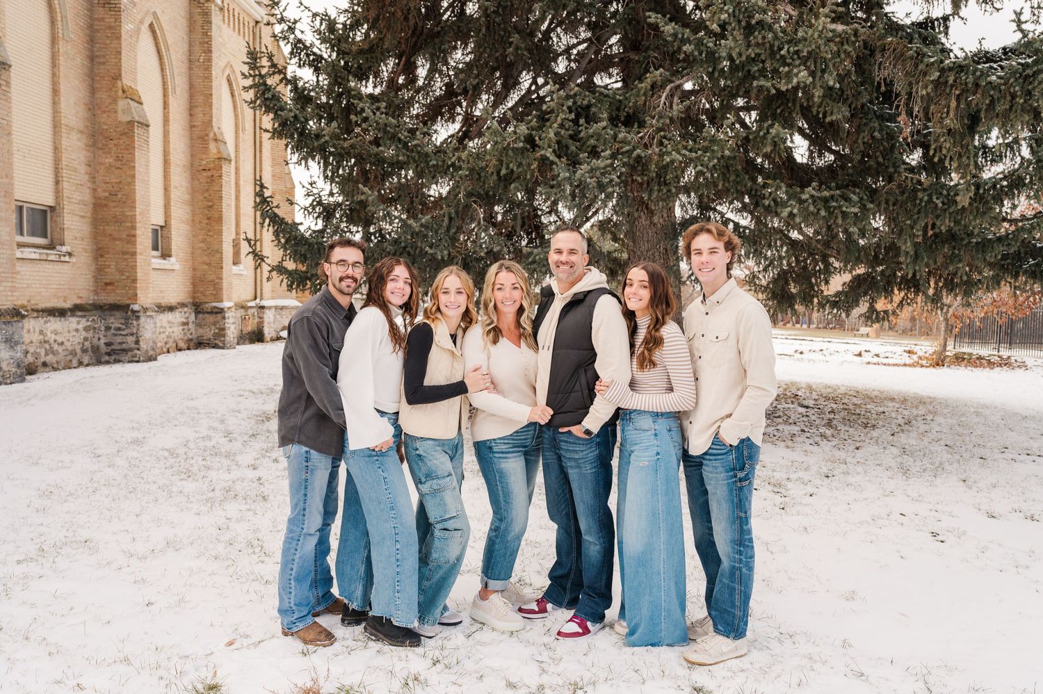 Group in coordinated denim and cream outfits posing in snow near stone building.