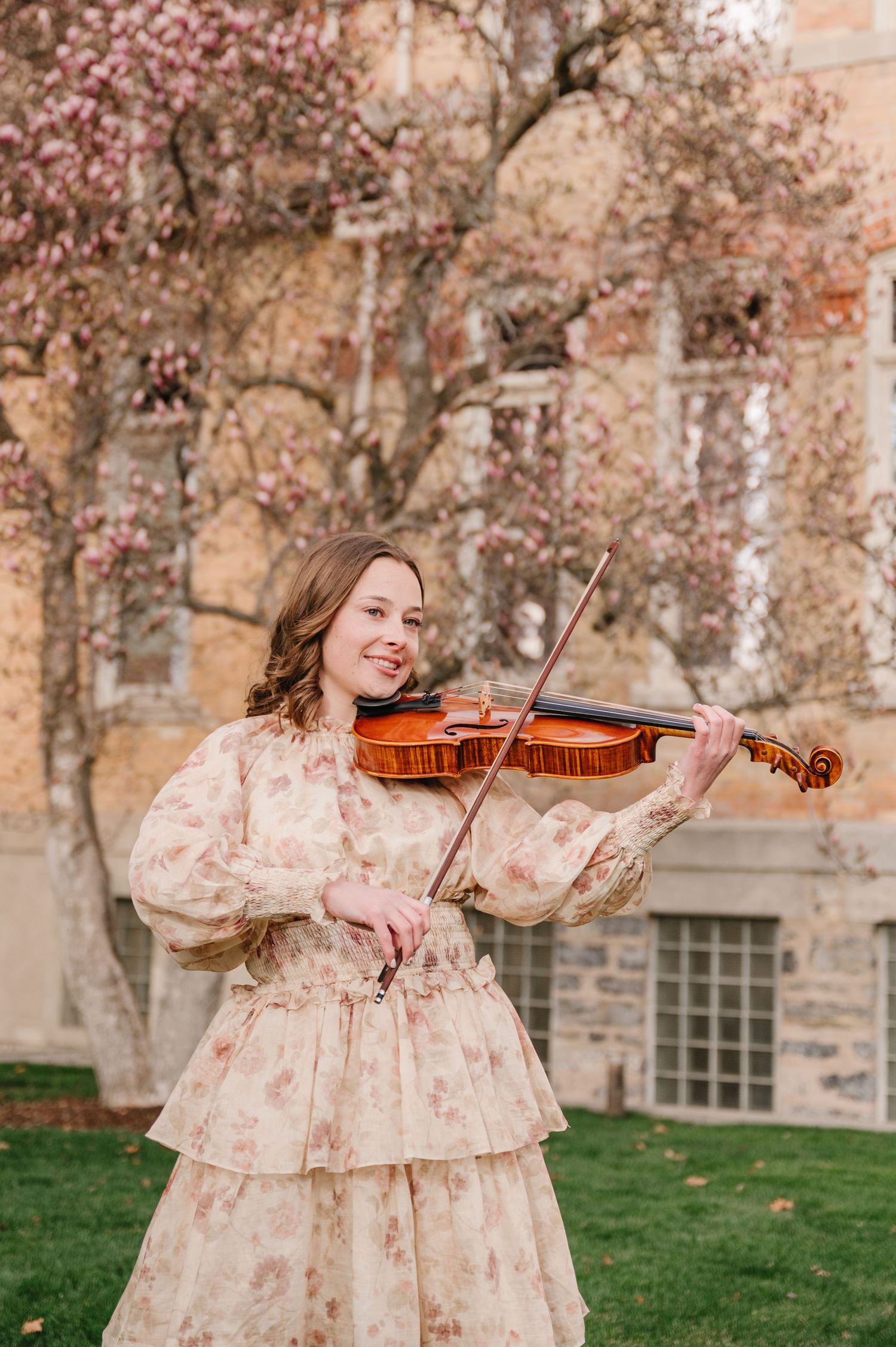 Musician plays violin outdoors in vintage floral dress with flowering tree and stone building in background.