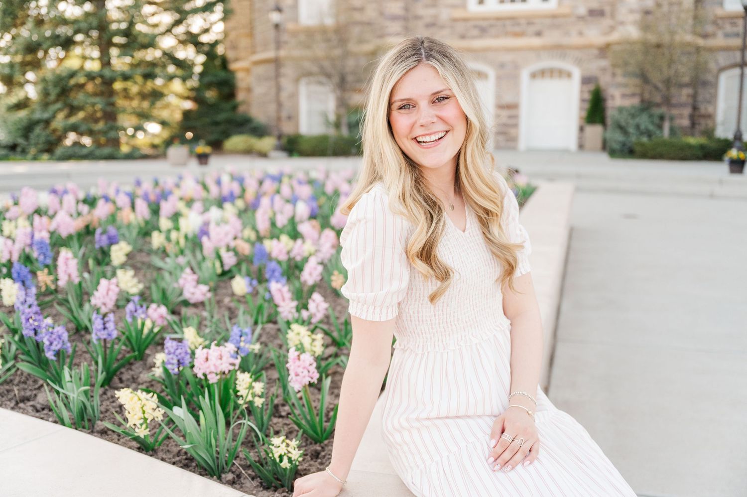 Person in white dress poses amid blooming spring flowers on campus grounds.