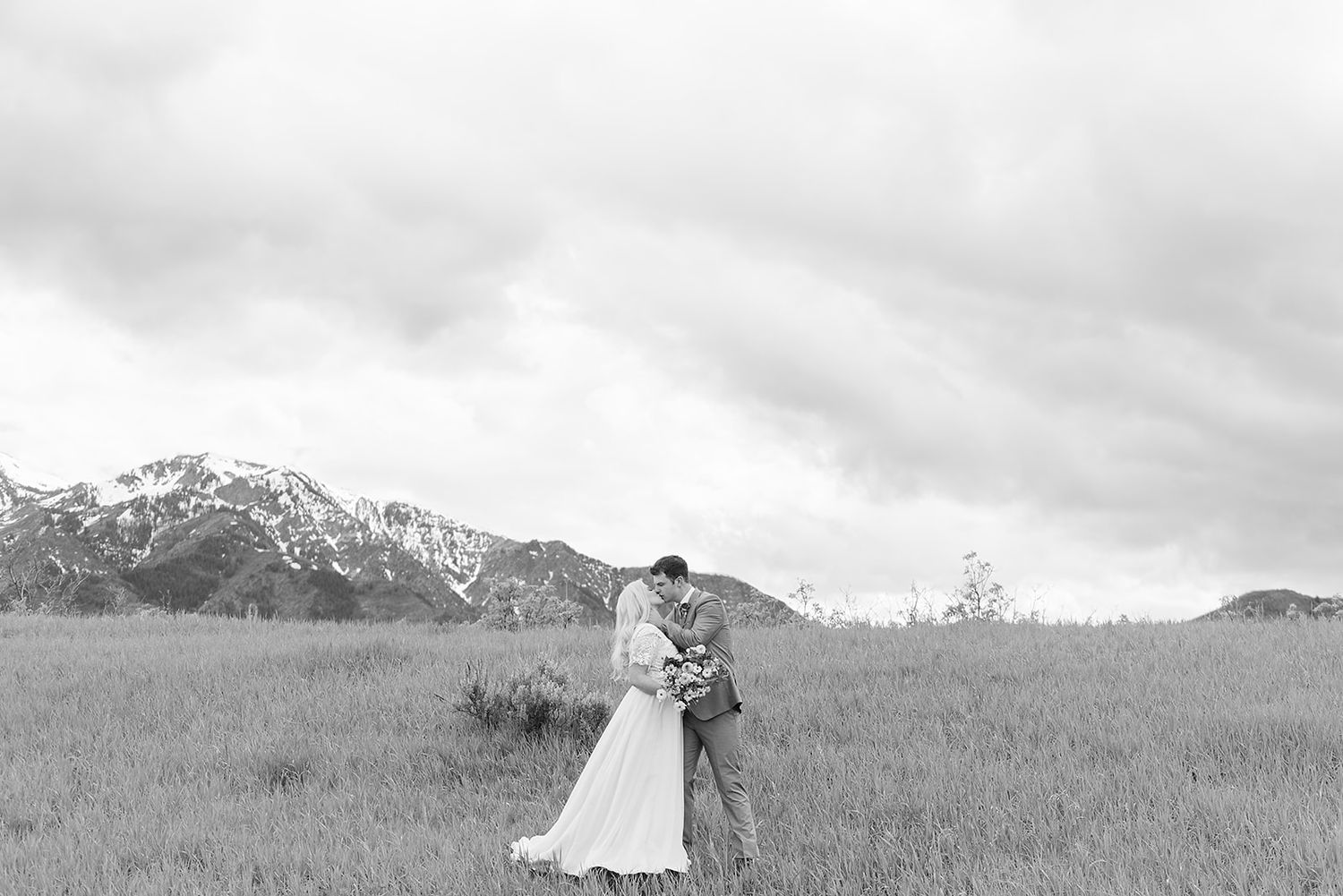 Black and white portrait of a couple embracing in a field with mountains in the background.