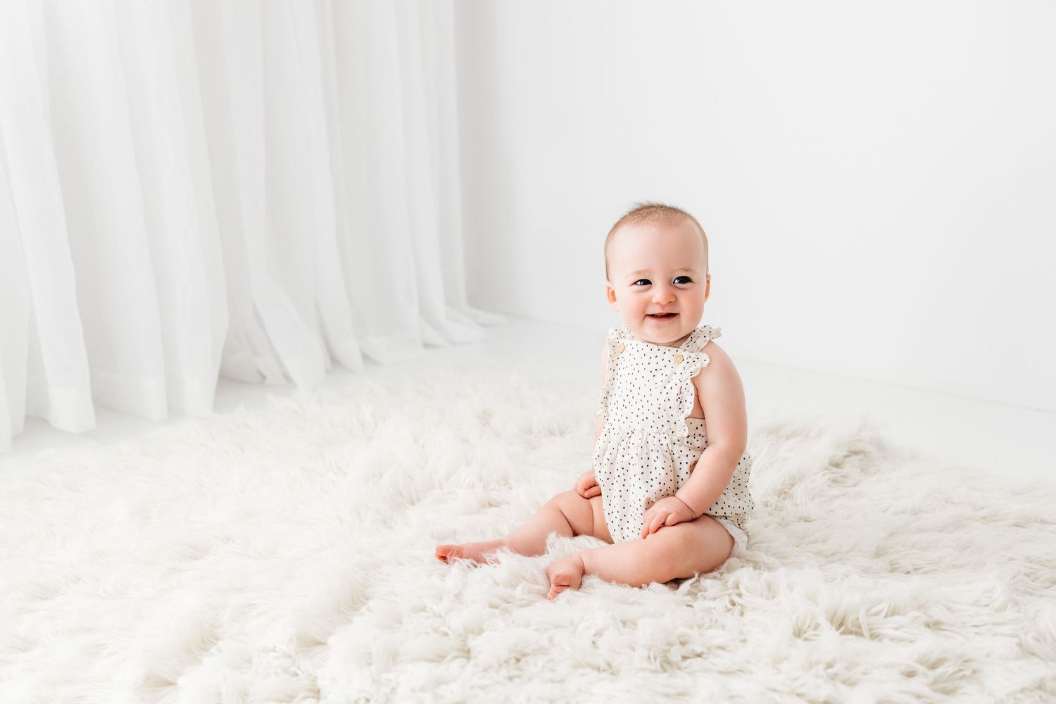 Baby wearing white lace romper sits on soft white fur rug against white curtain backdrop.