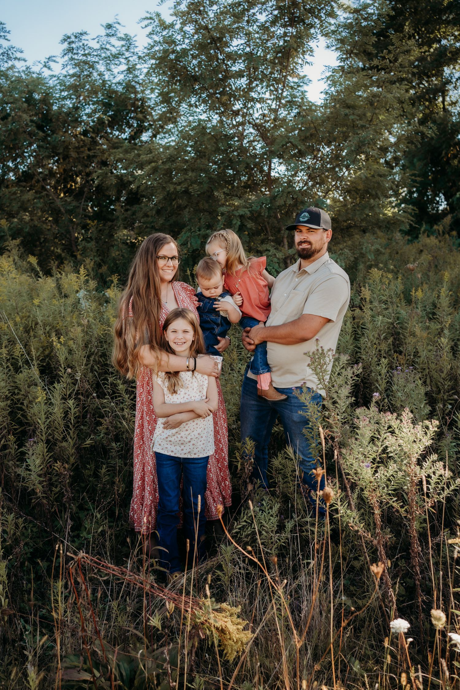 Family of four posing outdoors in a lush green field, parents with two young children smiling during a golden hour photo session.