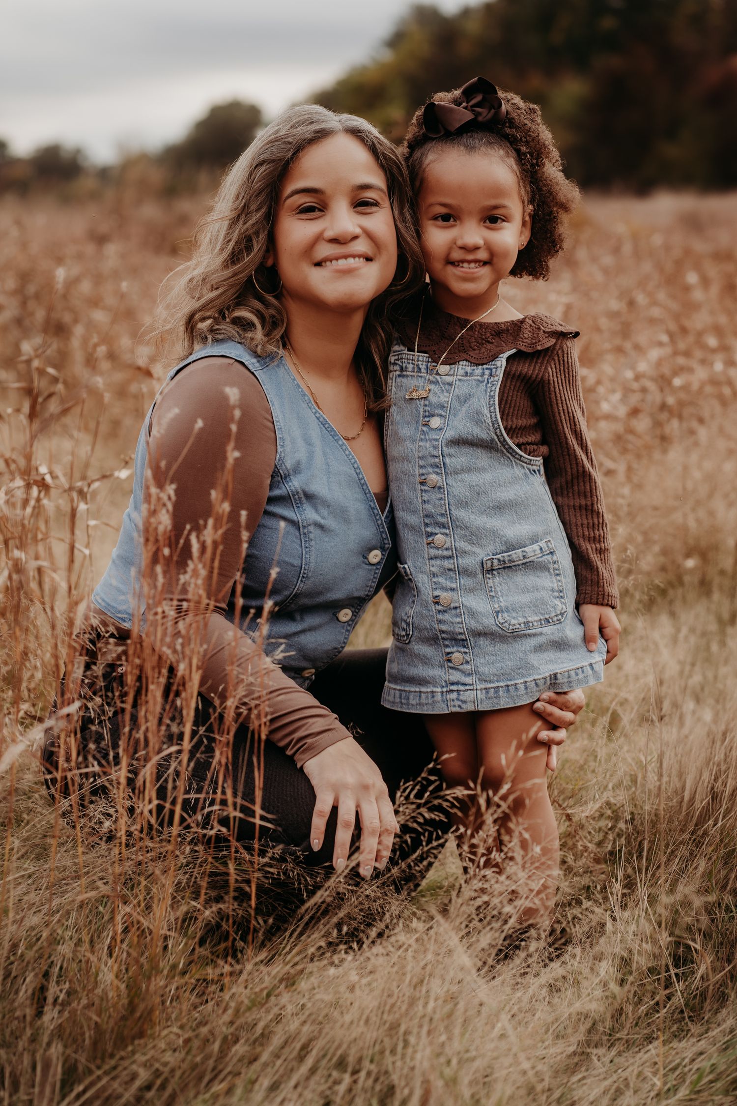 A woman and young girl in matching denim outfits smile together in an autumn field.