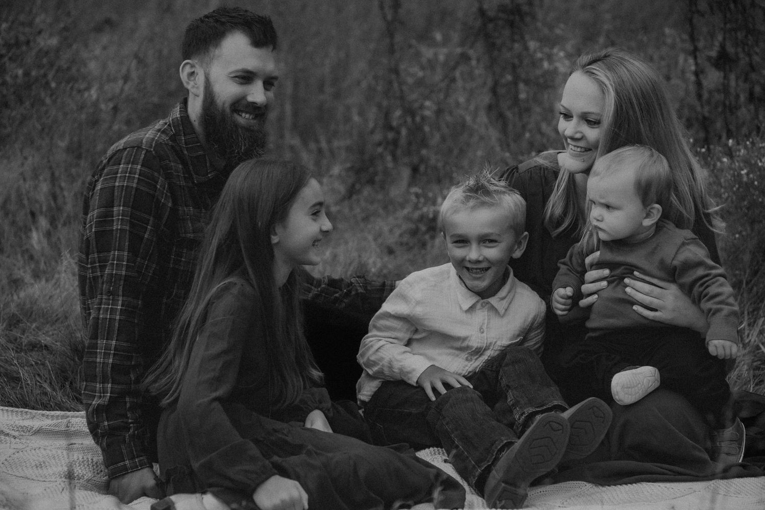 Black and white family portrait of parents with three children sitting outdoors, smiling and laughing together.