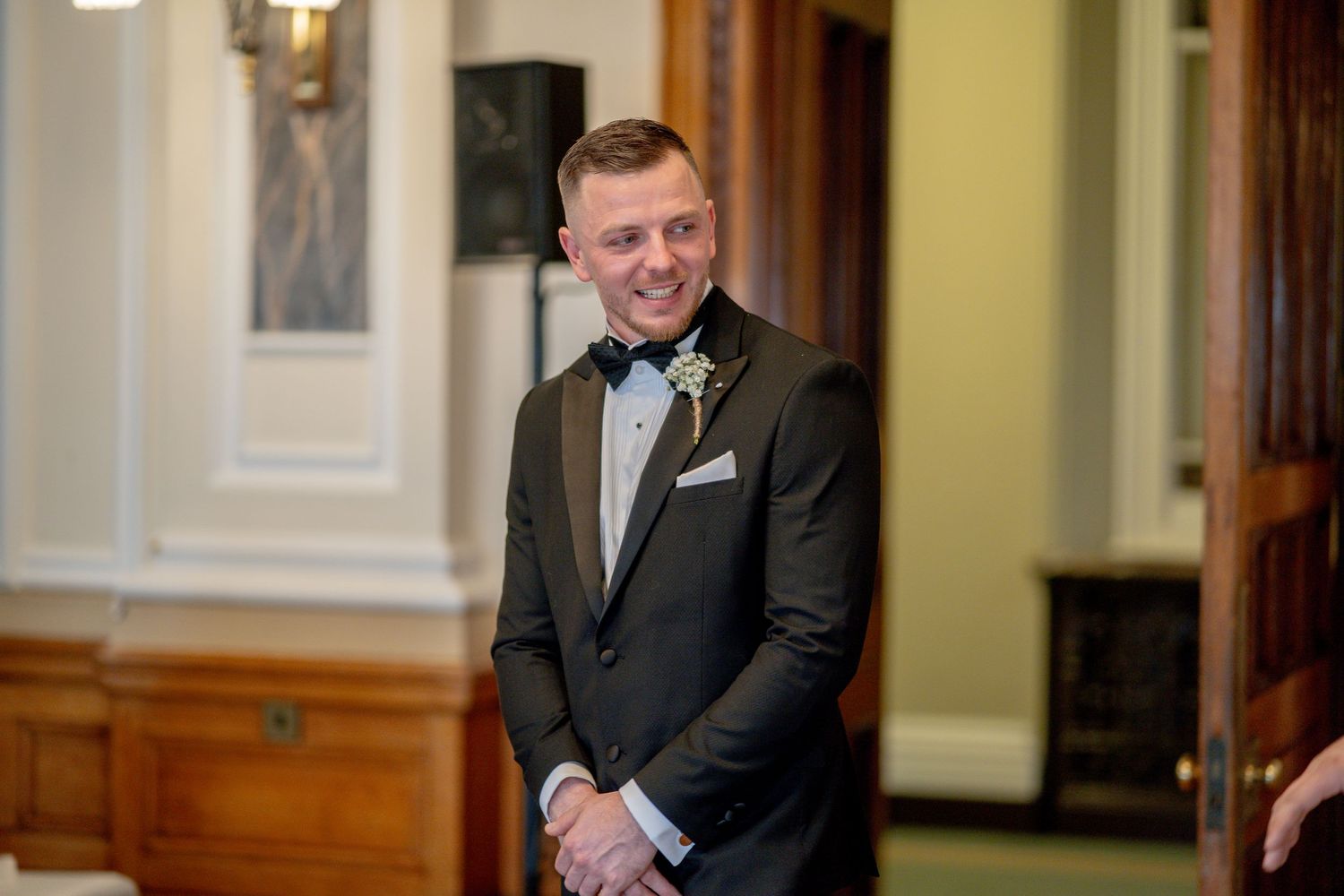 A groom in a black tuxedo and bowtie stands smiling in an elegant hallway with wooden paneling and columns.
