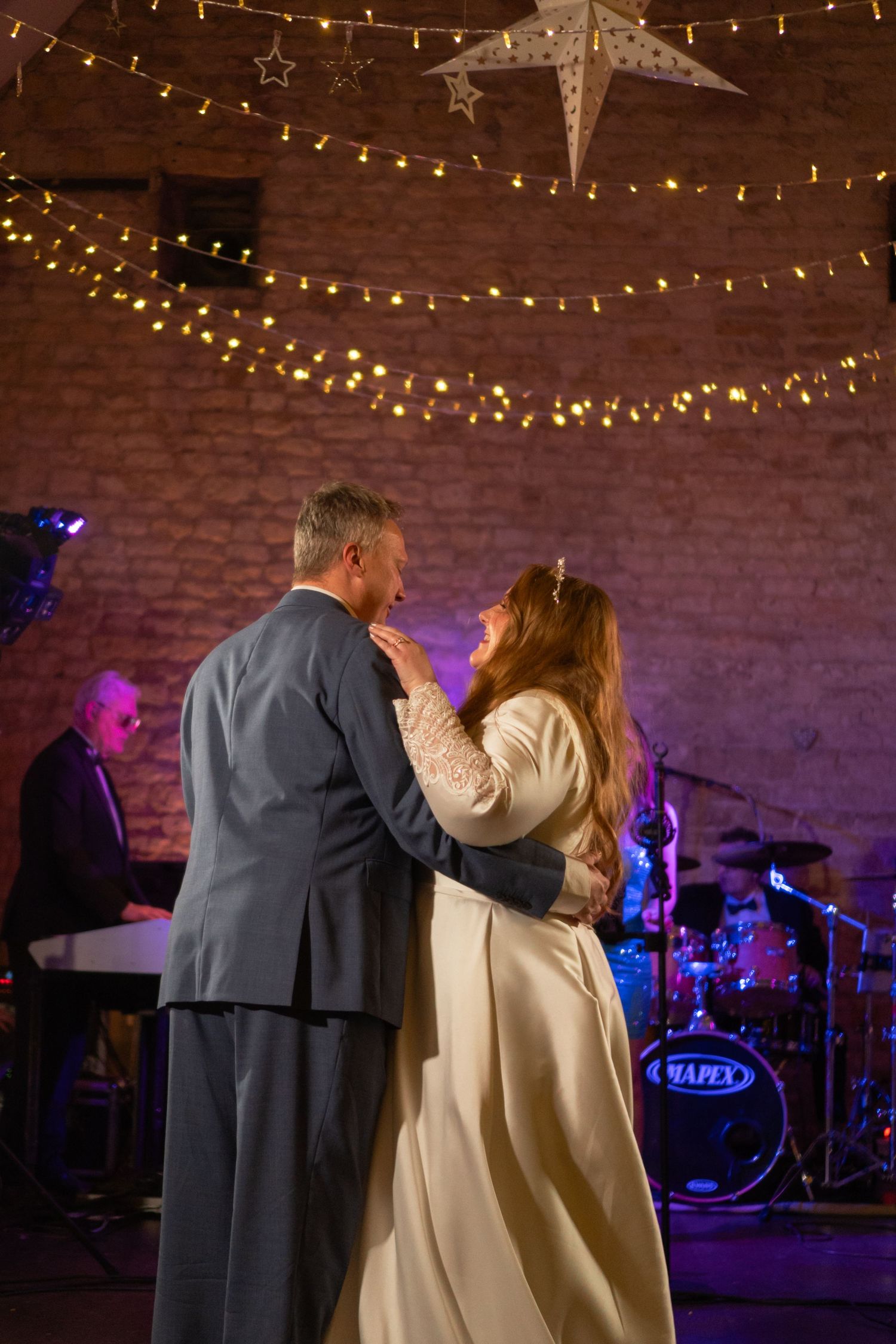 A couple dances at a wedding reception under string lights with a live band playing on stage behind them.
