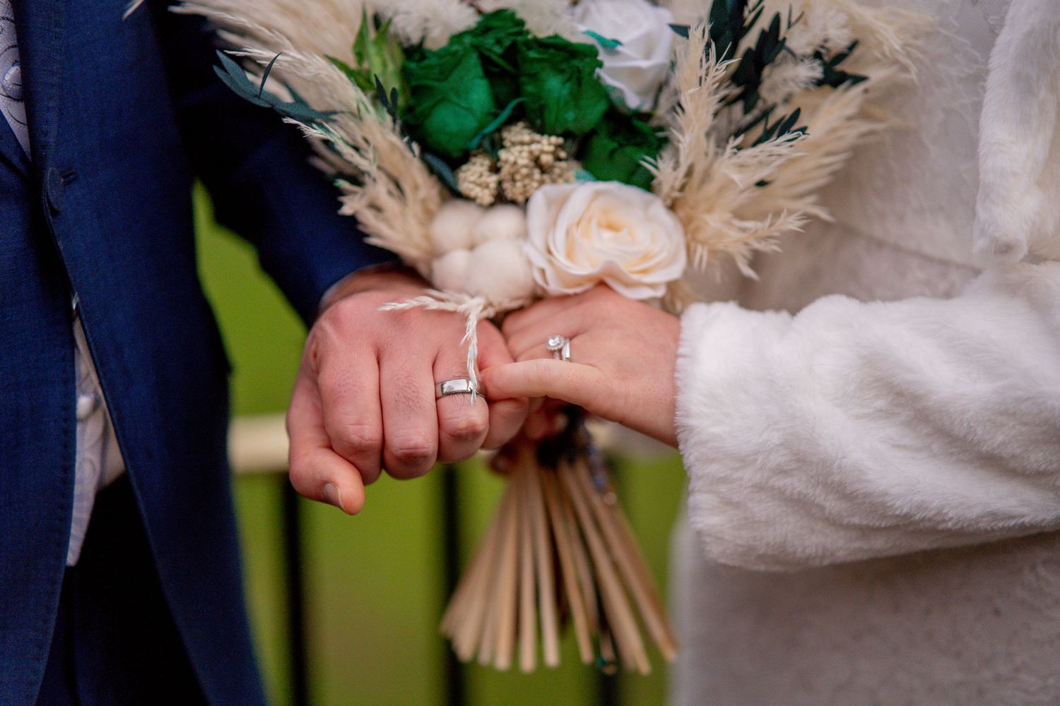 Close-up shot of hands exchanging wedding rings with a romantic bouquet featuring pampas grass and blush roses.