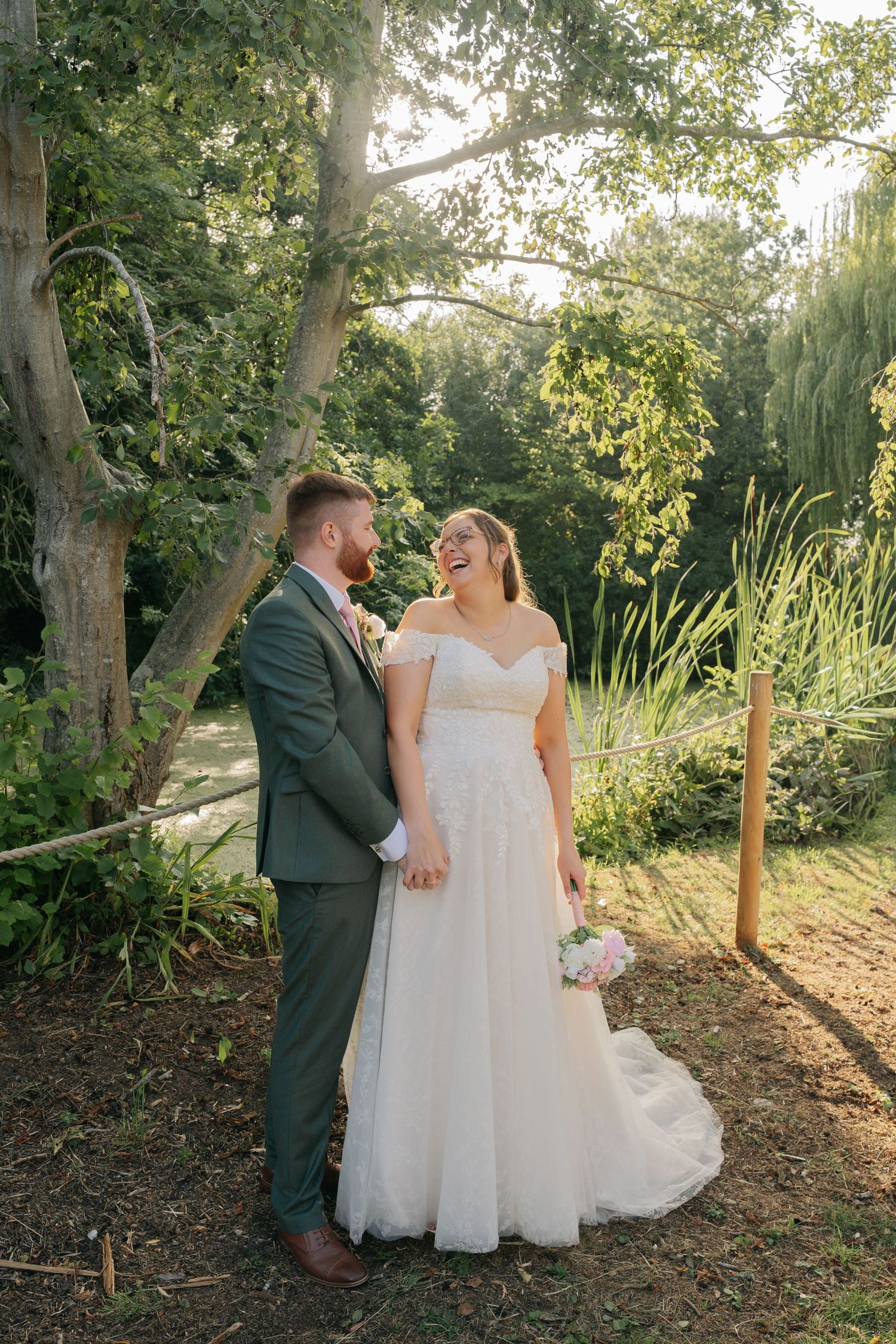 A couple in wedding attire stands together in a garden setting with sunlight filtering through trees.