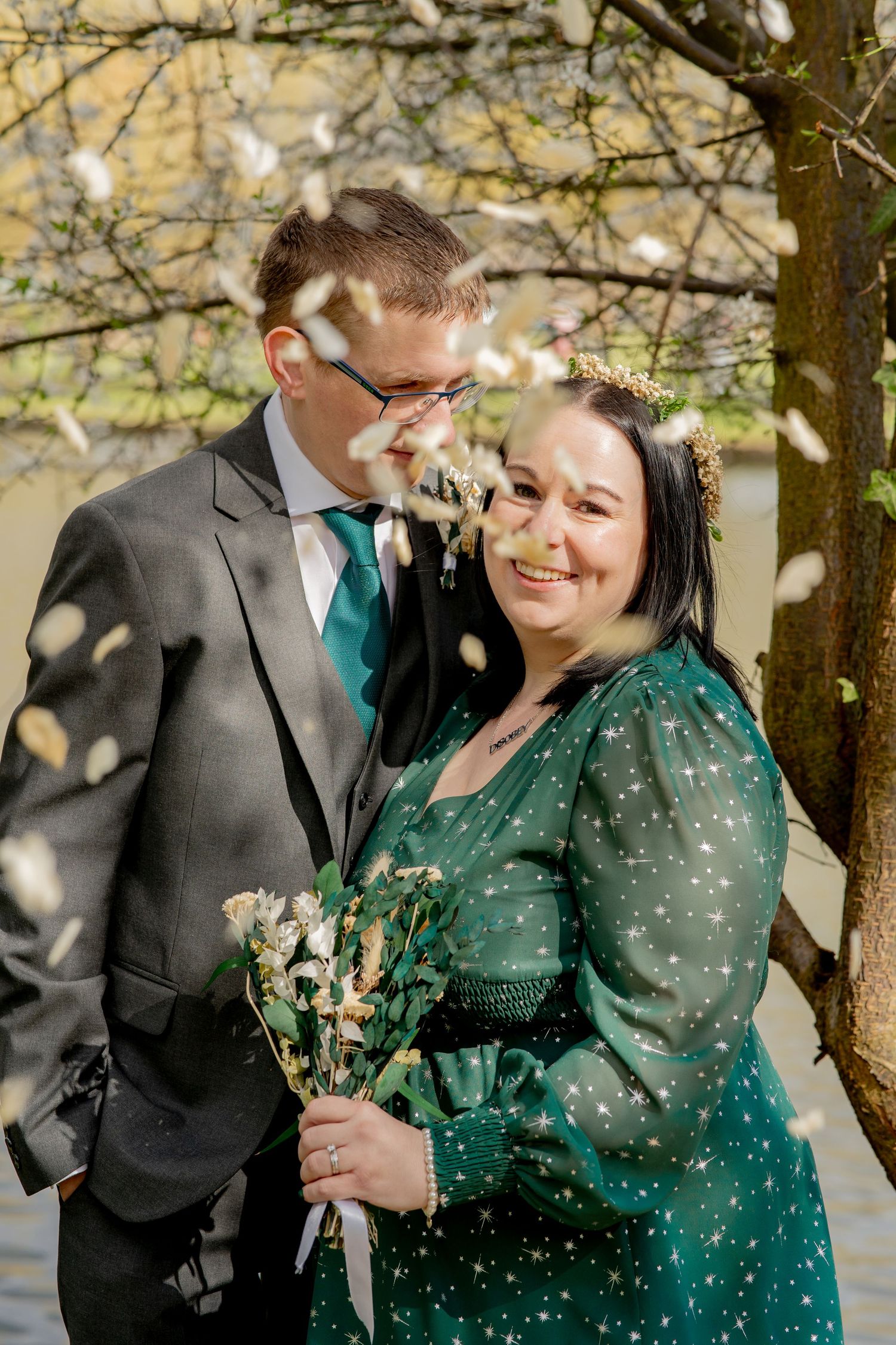 Happy couple sharing a joyful moment under spring blossoms, wearing coordinated green and grey formal attire.
